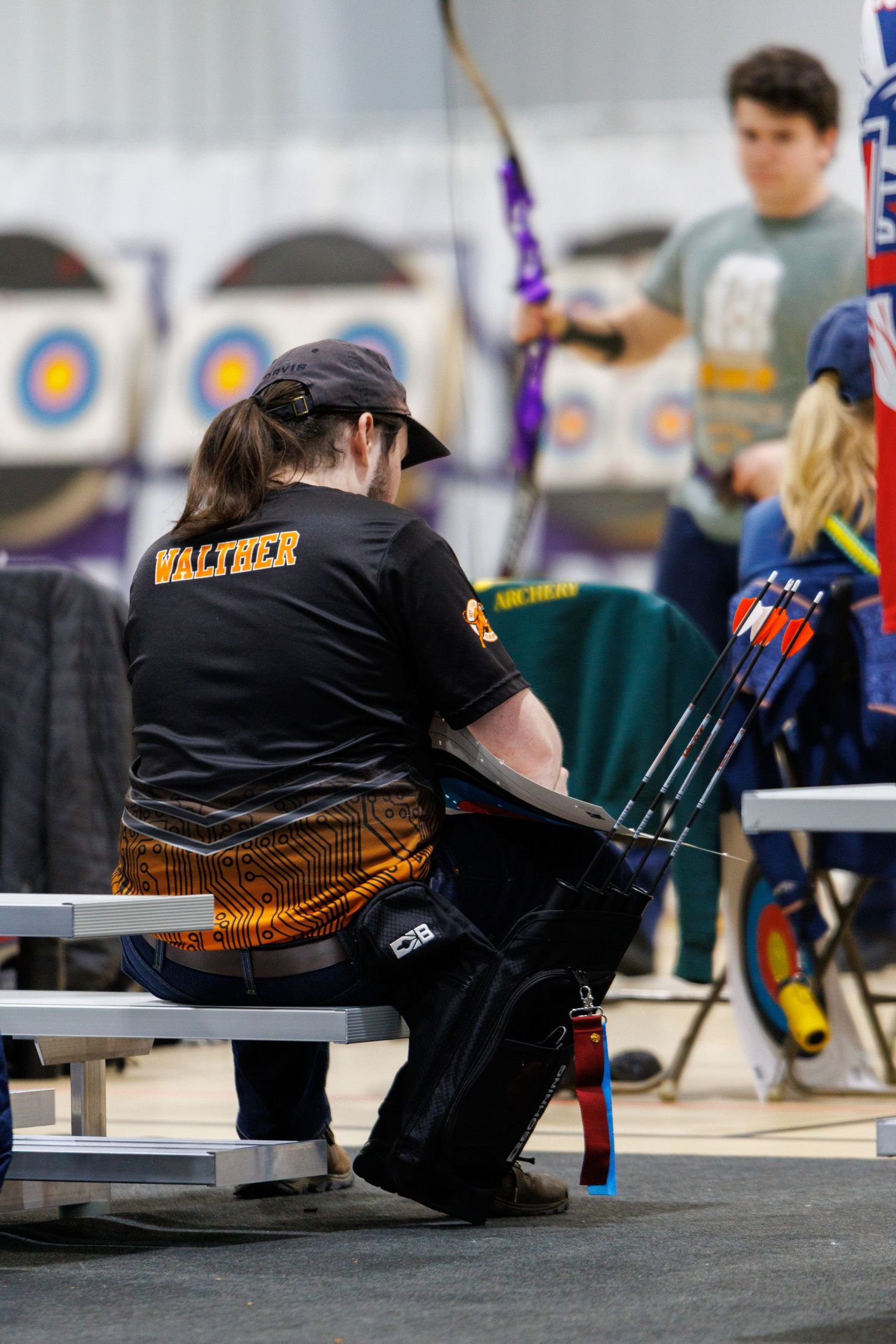 Archer wearing Walther jersey sitting on bench with arrows at an indoor archery tournament.