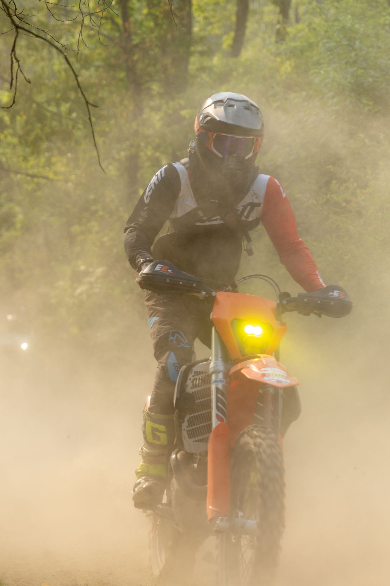 Multiple dirt bike riders kick up dust on an off-road trail while racing with orange and yellow lights visible.