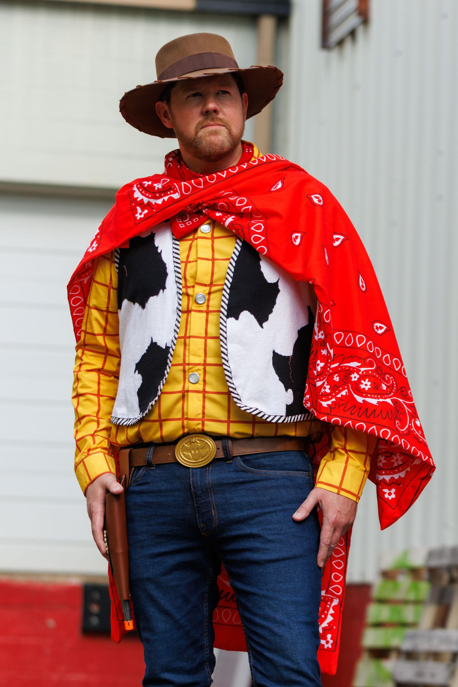 Man in a Woody-style cowboy costume with brown hat, red bandana cape, yellow plaid shirt and cow-print vest, holding a toy revolver.