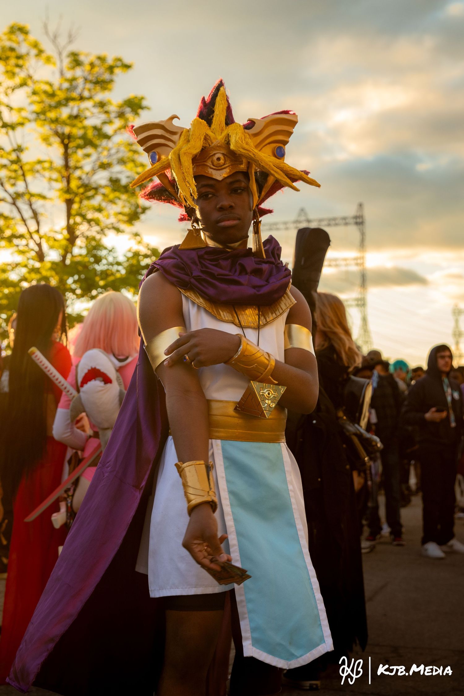 Cosplayer wearing an ornate gold crown and purple cape poses at sunset during an outdoor convention.