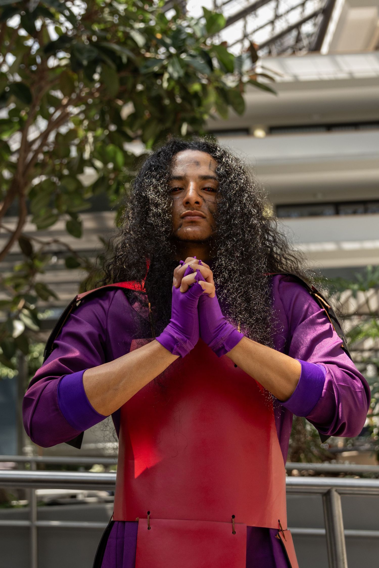 A person with long curly dark hair wearing purple gloves and a red and purple outfit poses with clasped hands in prayer.