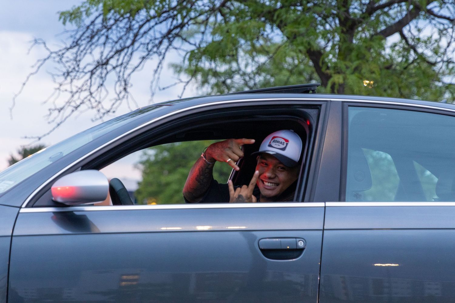 A person sits in a grey car wearing a baseball cap and gives a thumbs up gesture through the window.