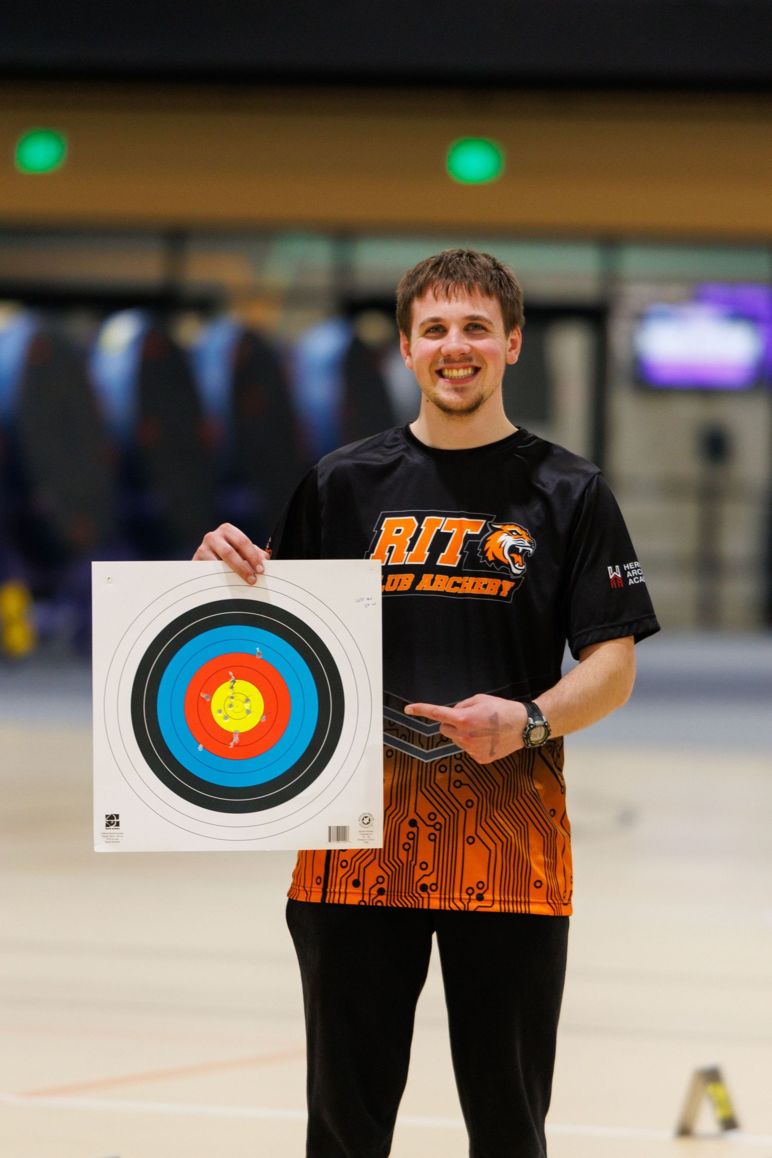 RIT club archery member smiling and holding a colorful target at an indoor archery competition.