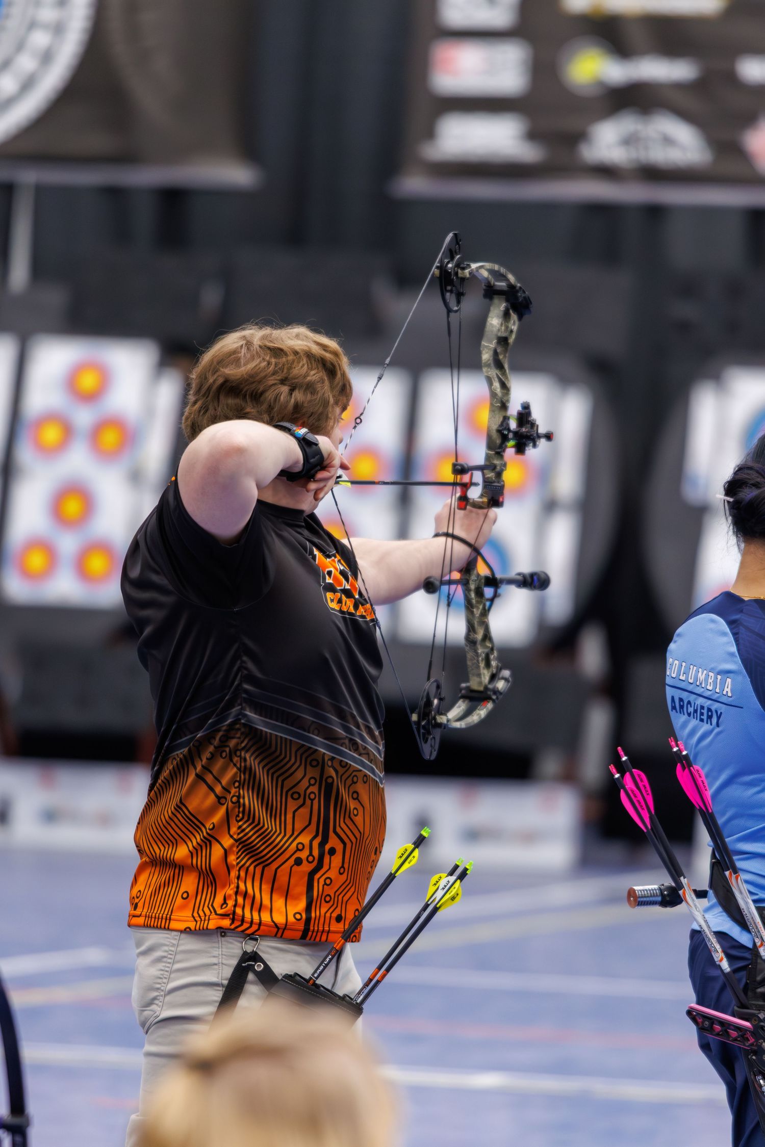 Archer aiming compound bow at indoor archery range, wearing orange circuit-board jersey, with targets visible in background.