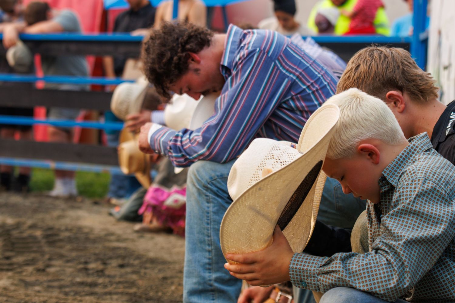 Two boys in cowboy hats and plaid shirts sit along a rodeo fence, heads bowed during an event.