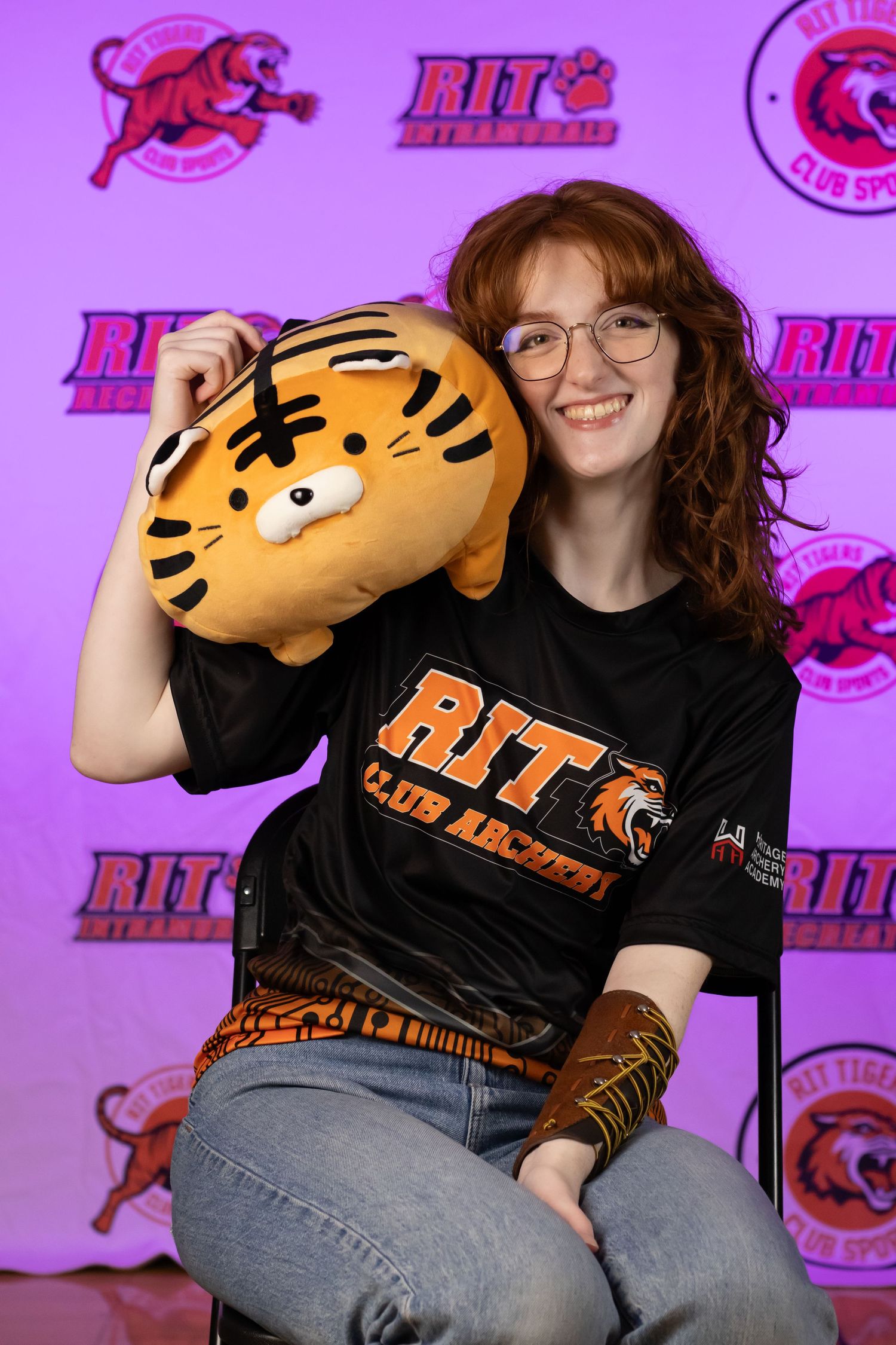 Student with RIT Club shirt holds plush tiger mascot against purple backdrop.