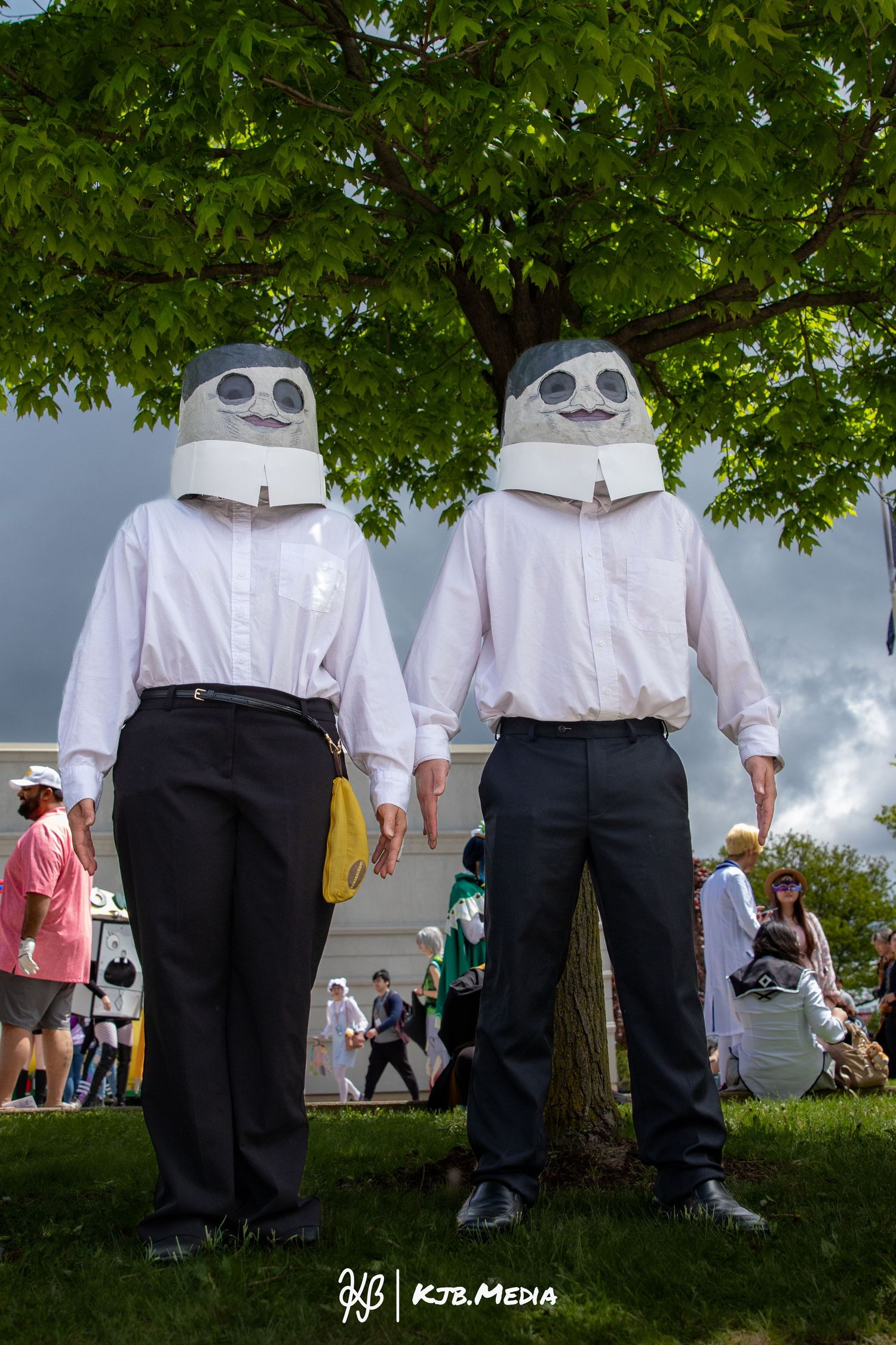 Two figures in matching white shirts and robot head masks pose under a green tree.
