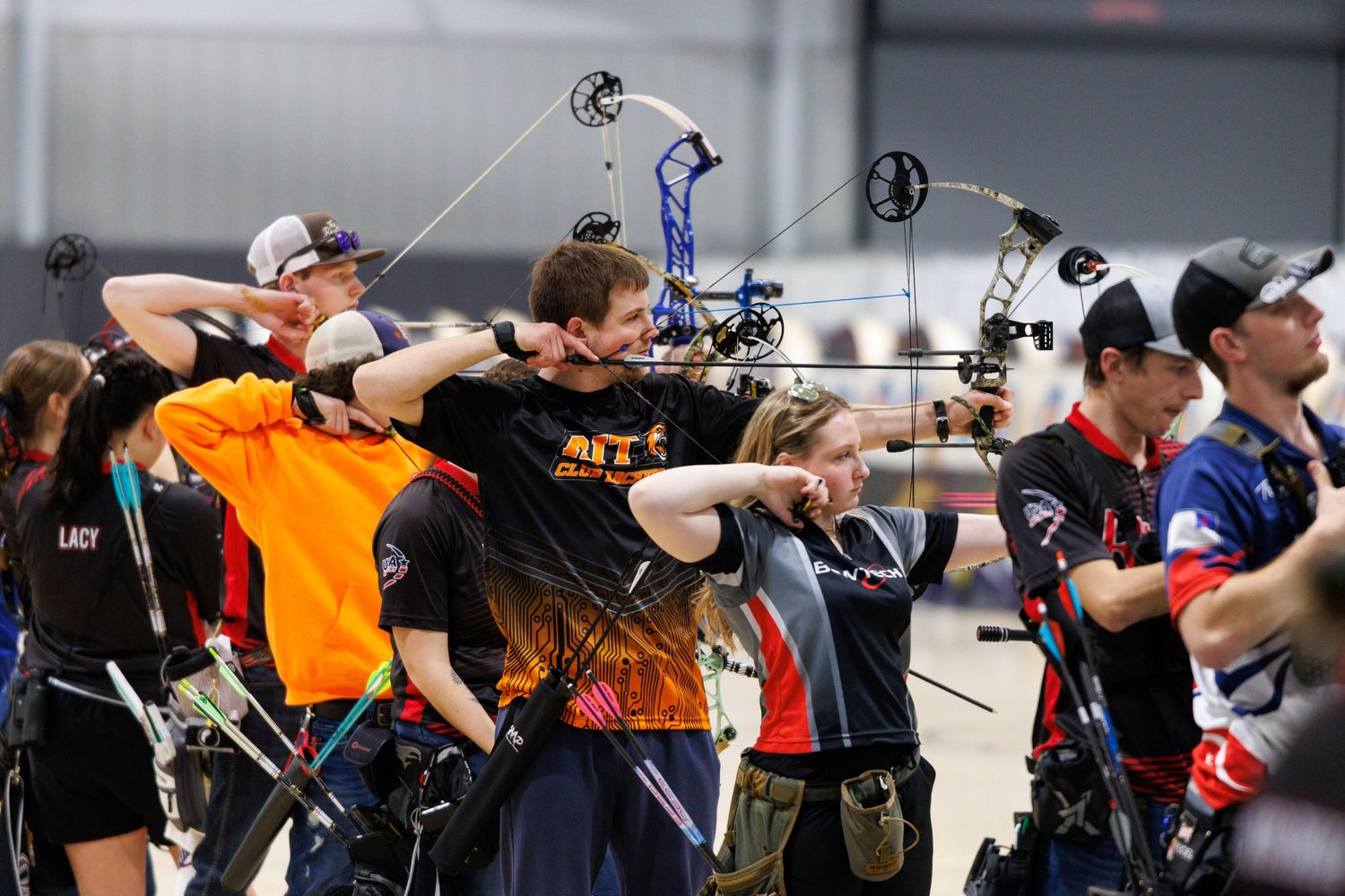 Group of archers draw compound bows simultaneously at an indoor archery competition.
