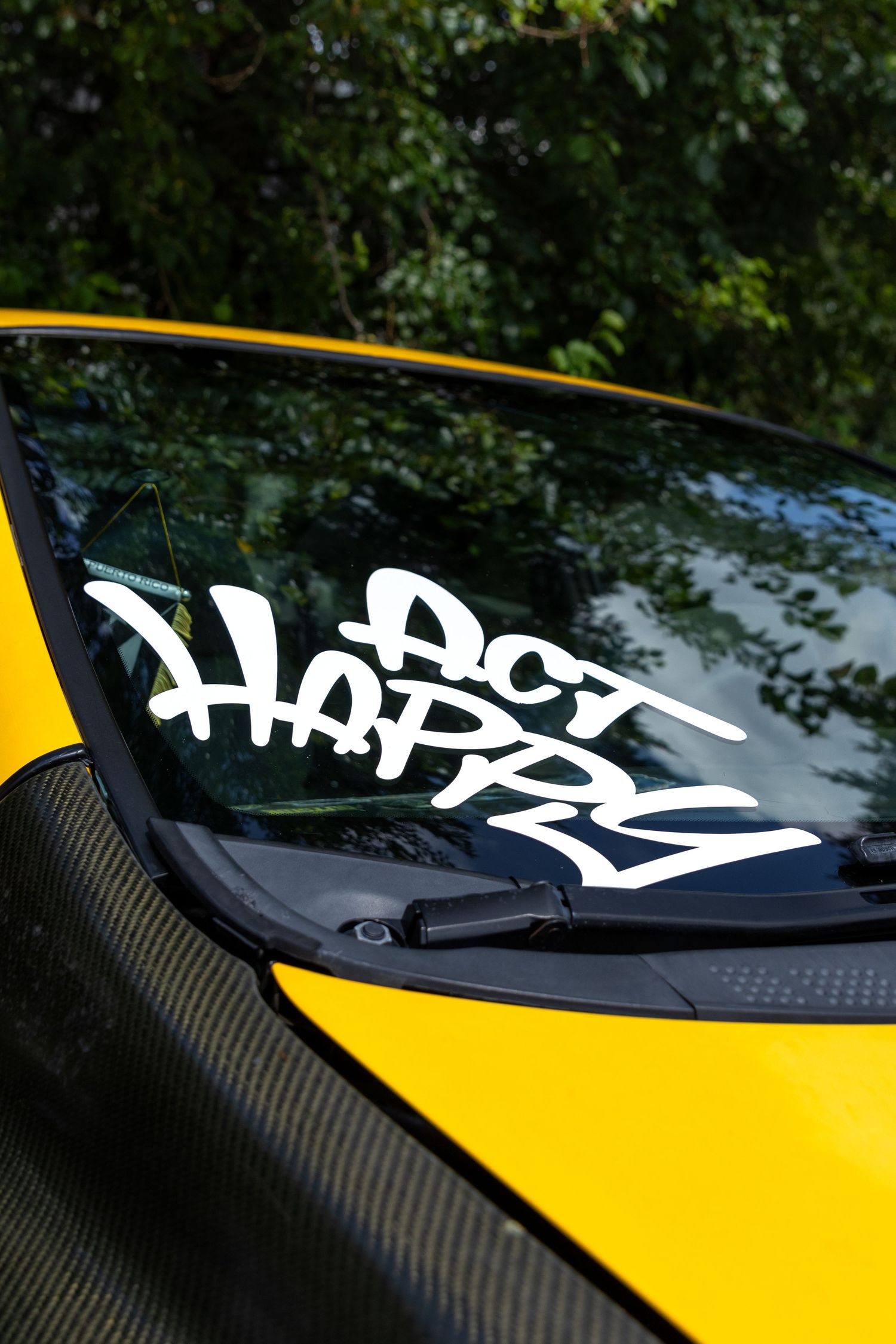 White graffiti style decal on yellow car windshield with carbon fiber detail and green foliage reflected in glass.