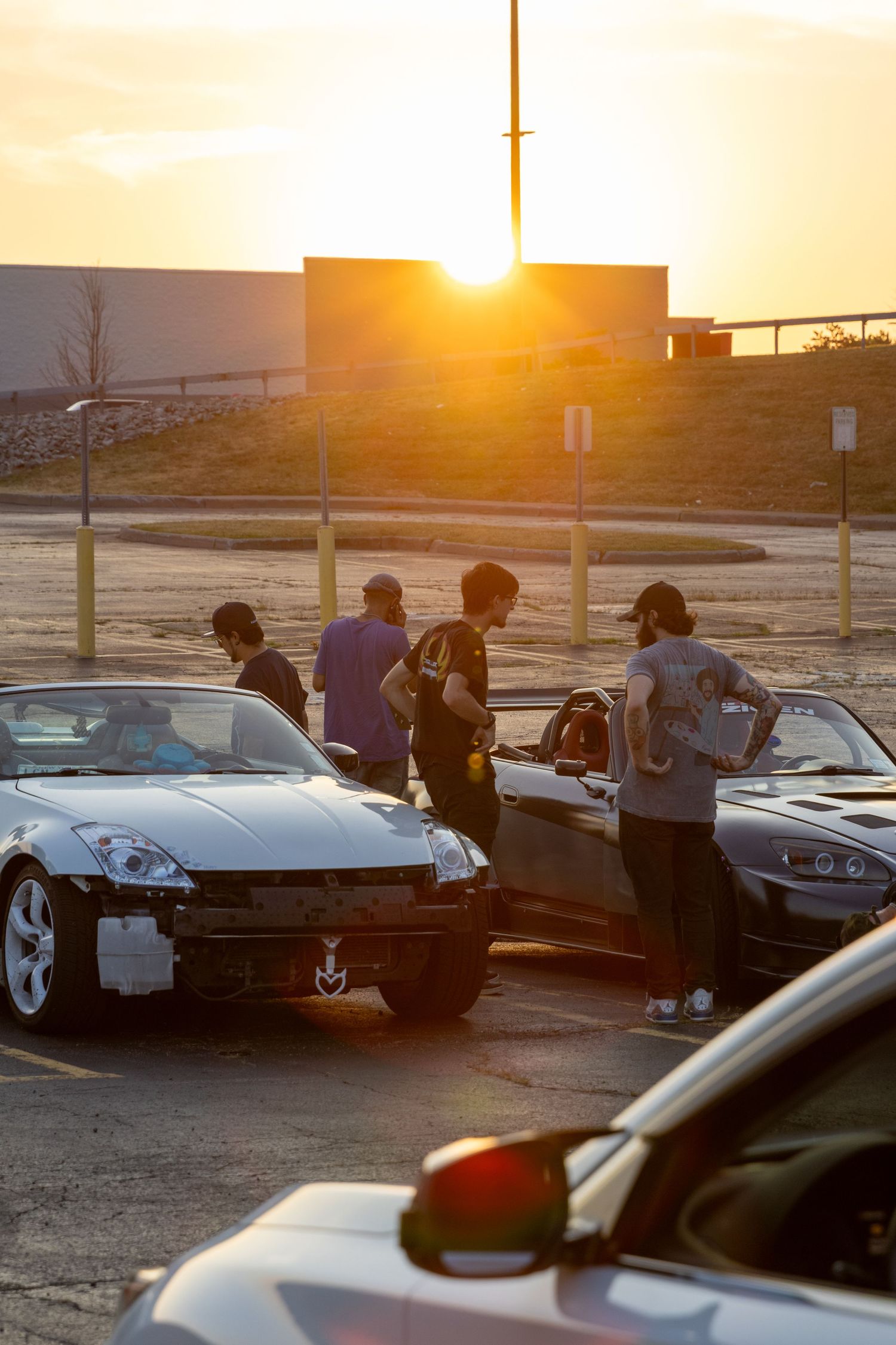 A group of car enthusiasts gather around modified sports cars in a parking lot during golden hour sunset.