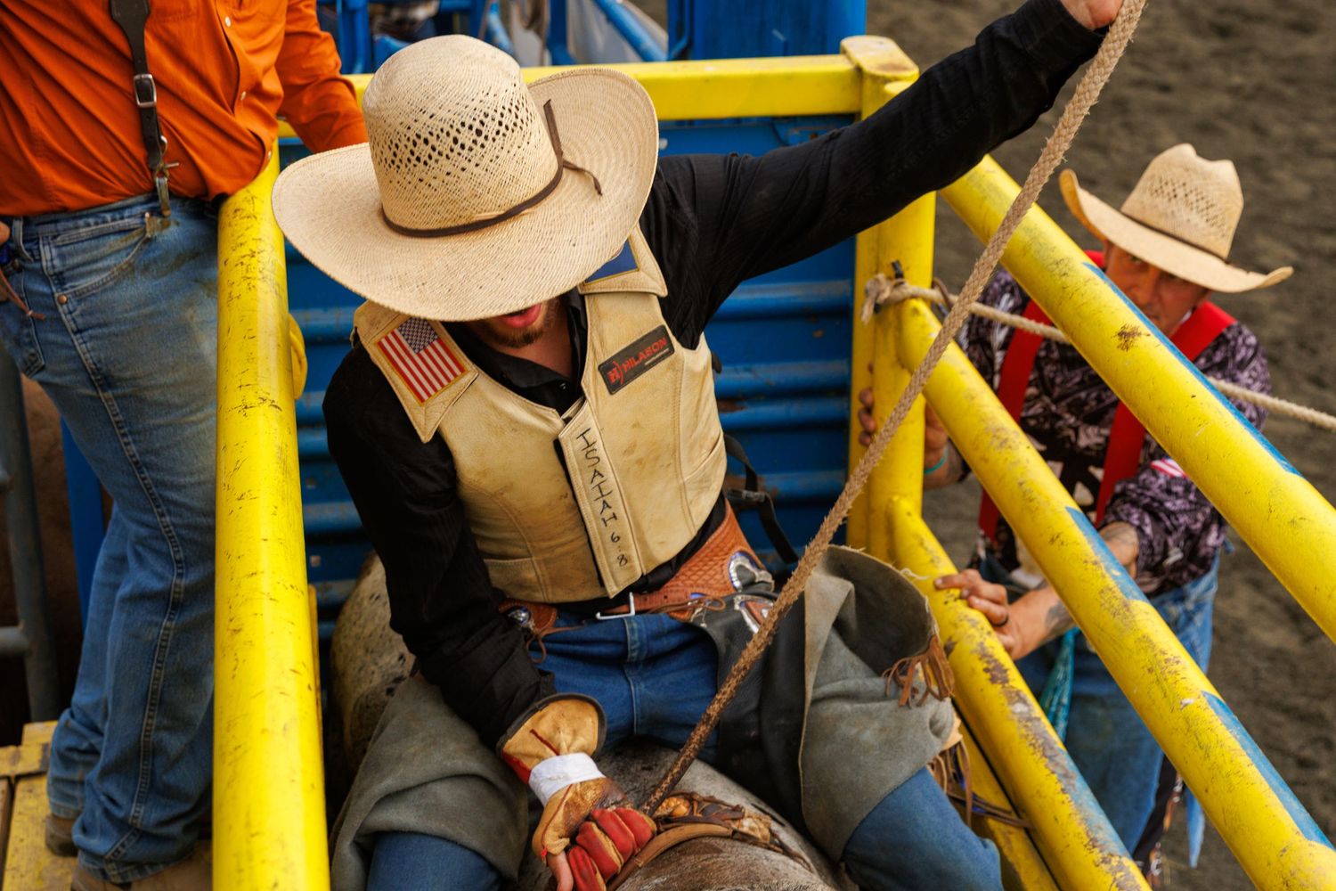 Cowboys prepare for a bull ride at a rodeo, adjusting gear behind yellow chutes in western attire.