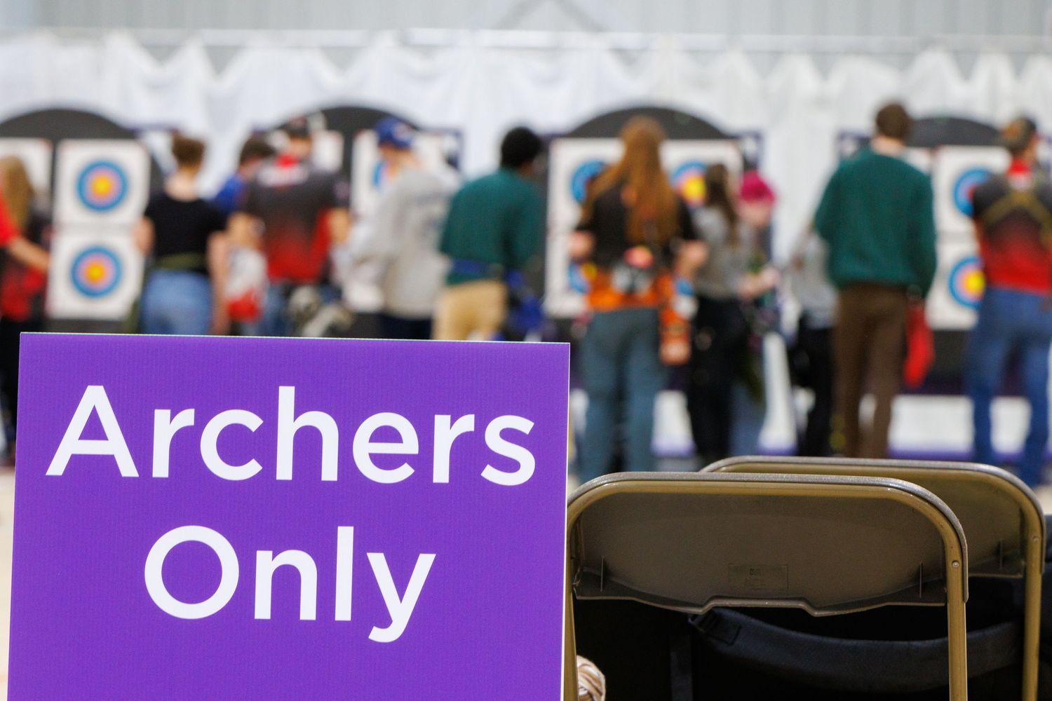 Purple Archers Only sign at an indoor archery tournament with targets and participants in the background.