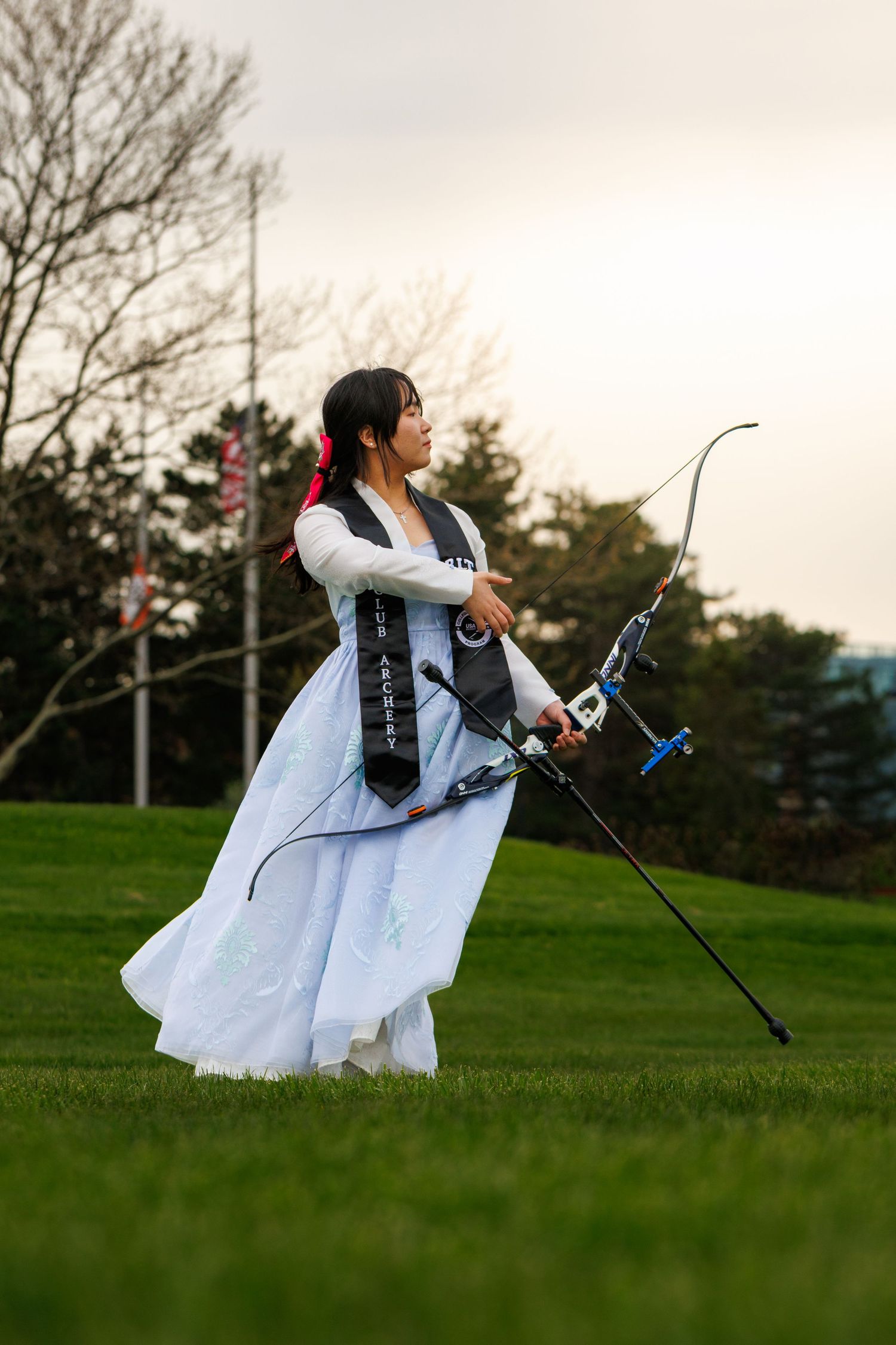 Woman in traditional white hanbok dress practicing archery with a recurve bow on a green lawn.