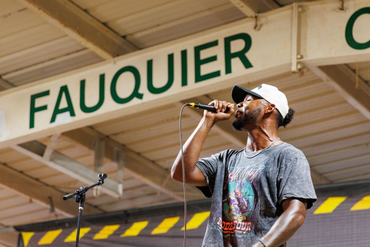 Man singing into microphone on stage at Fauquier County event, wearing white cap and gray t-shirt.