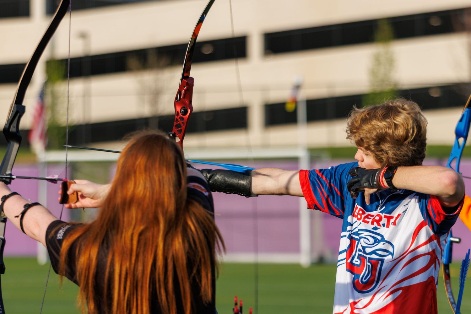 Two archers drawing bows at an outdoor archery range, one with red hair, competing in a recurve archery event.