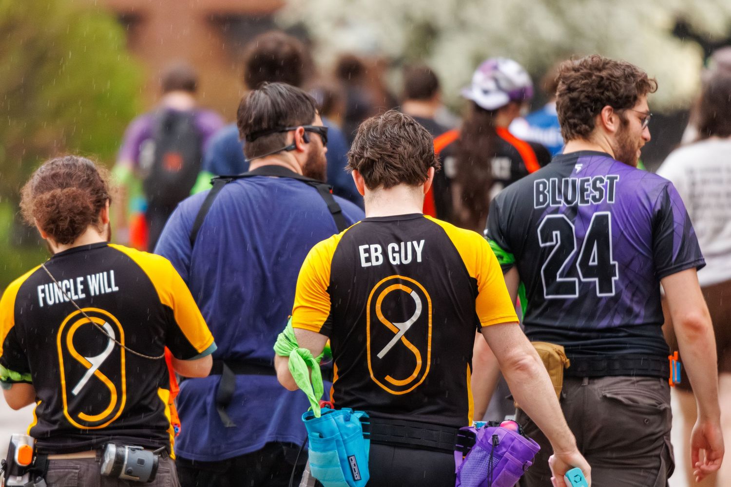 Group of players in colorful jerseys with nicknames like EB GUY and BLUEST 24 walking at an outdoor sports event.