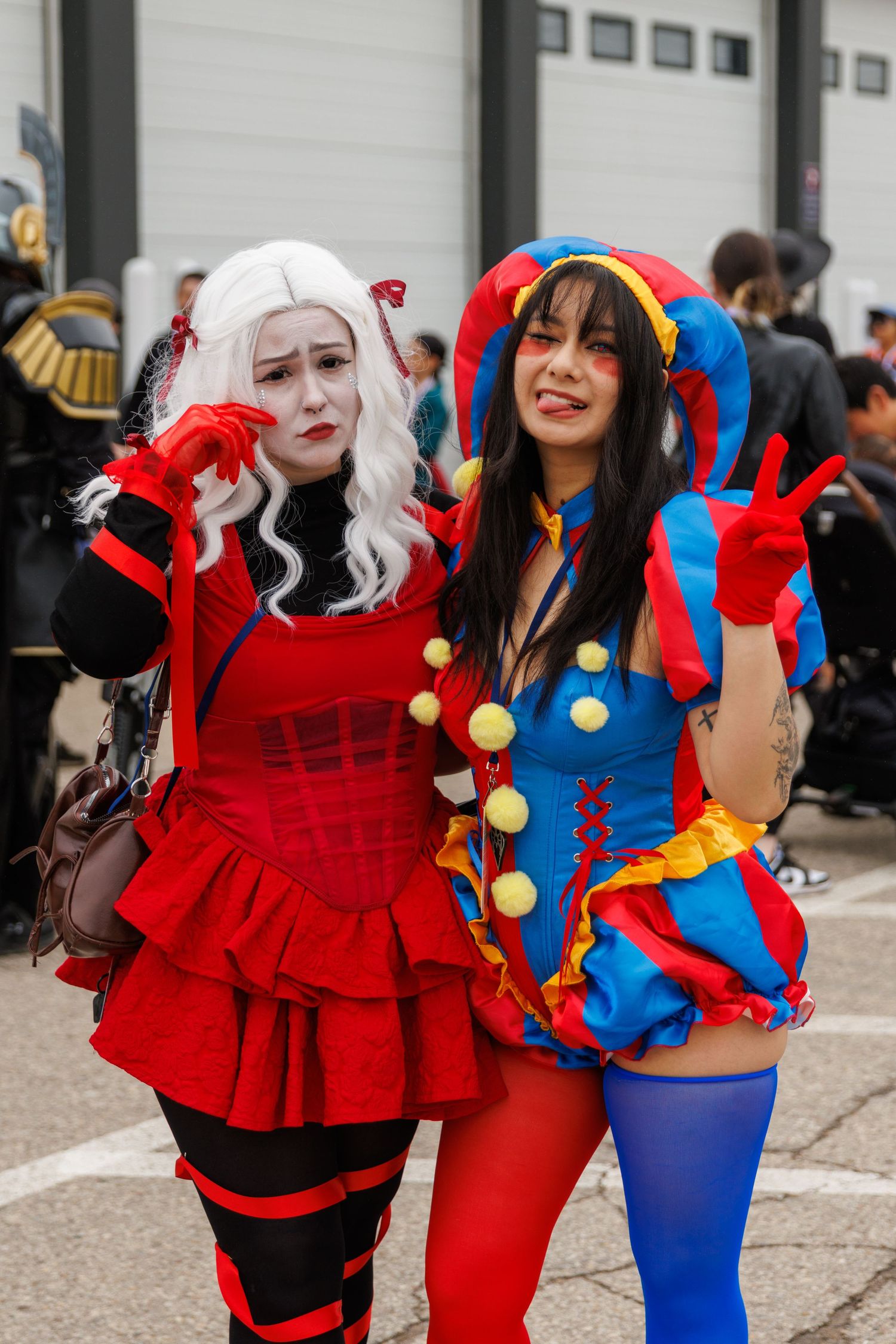 Two female cosplayers in colorful red and blue jester-inspired costumes pose together at an outdoor convention.