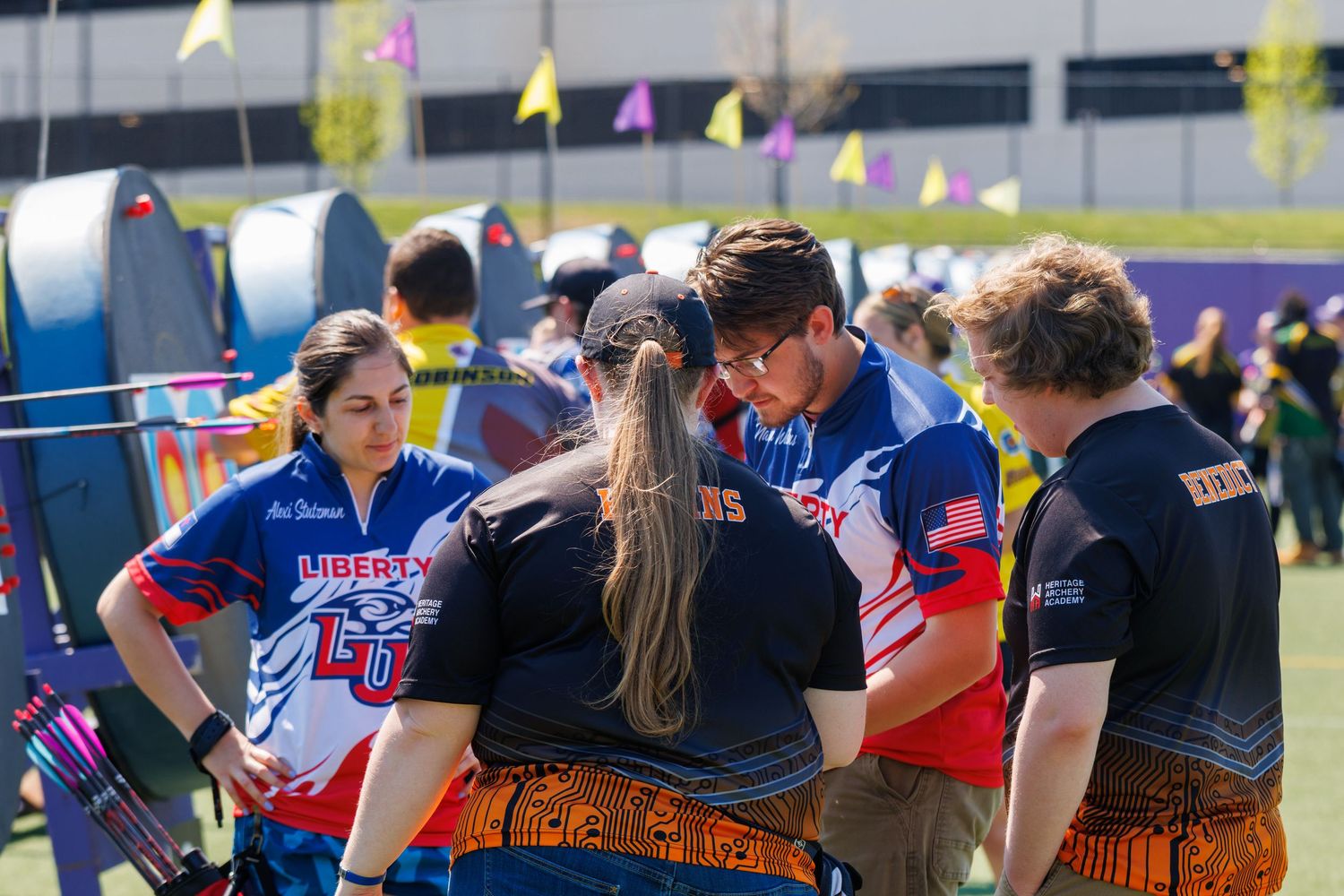A team of archers in blue uniforms huddle together during an outdoor archery tournament on a sunny day.