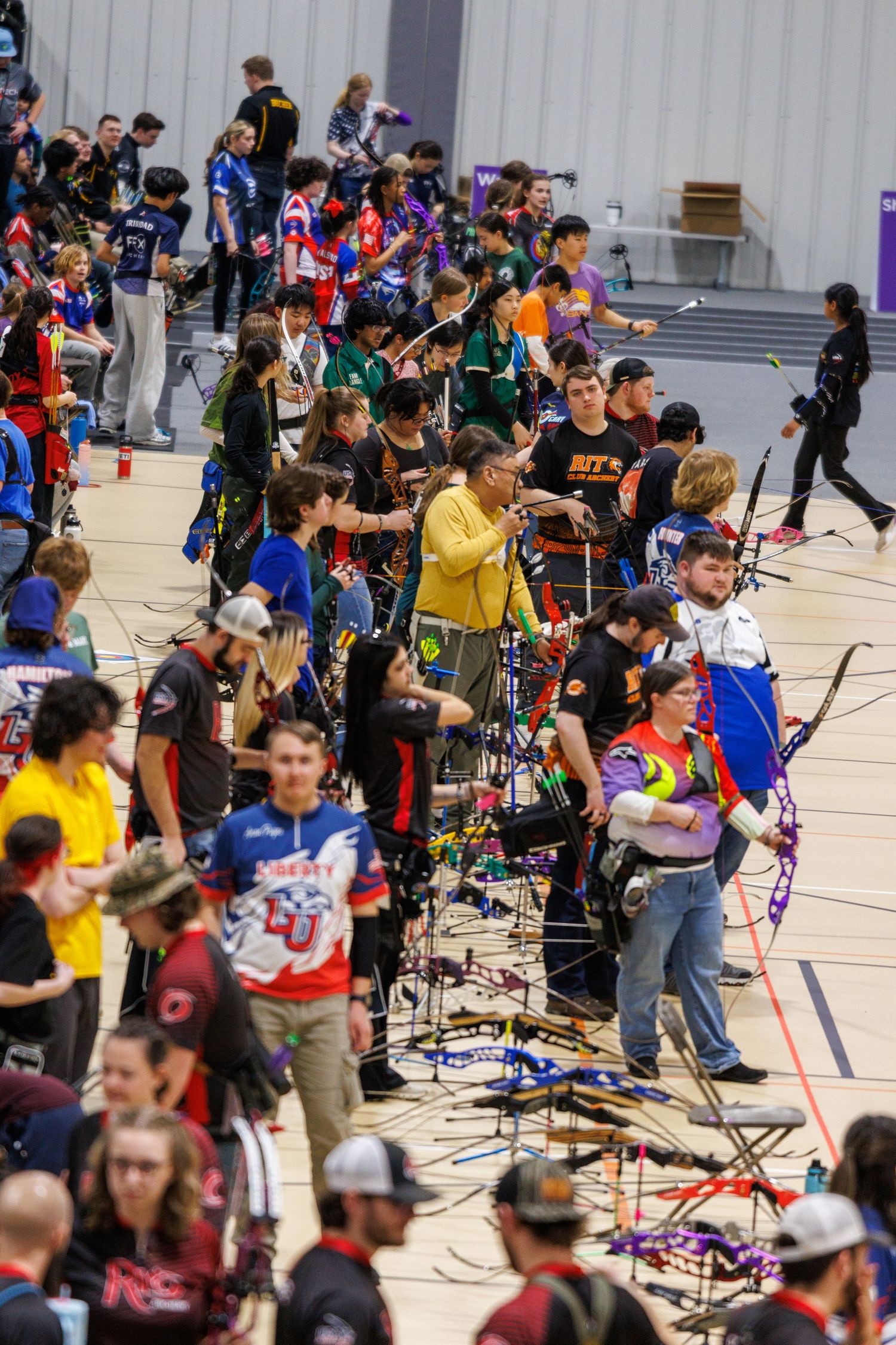 Large group of archers competing in a crowded indoor archery tournament with colorful bows and targets.
