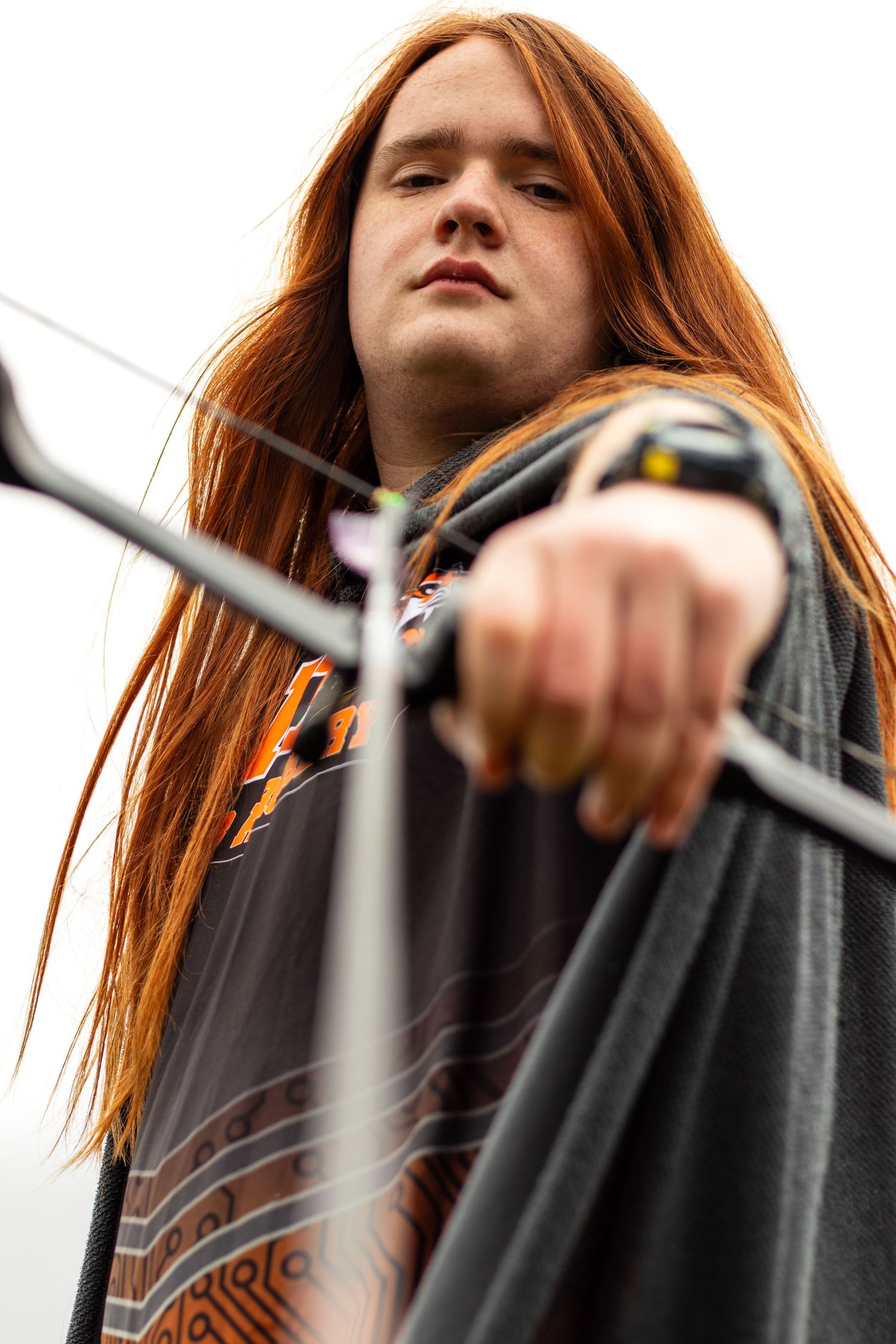 Low angle shot of archer with long red hair holding bow against grey sky.