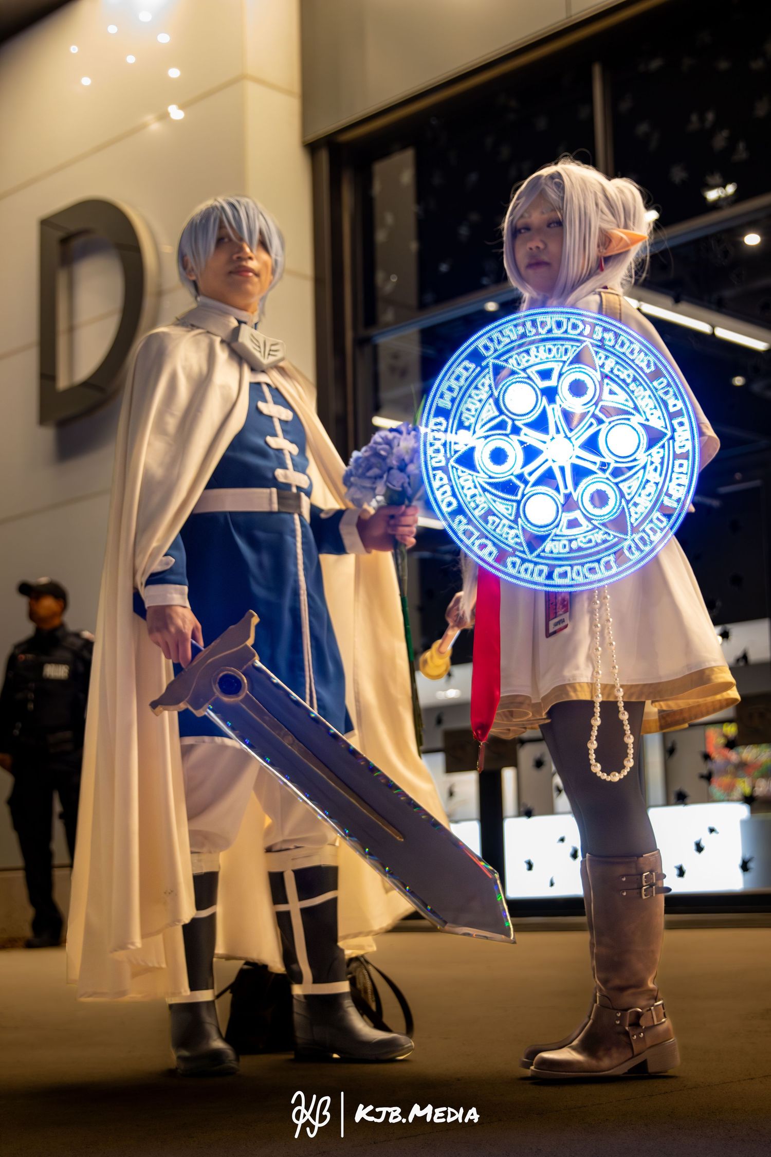 Two cosplayers in white and blue fantasy outfits, one holding a glowing magical shield.