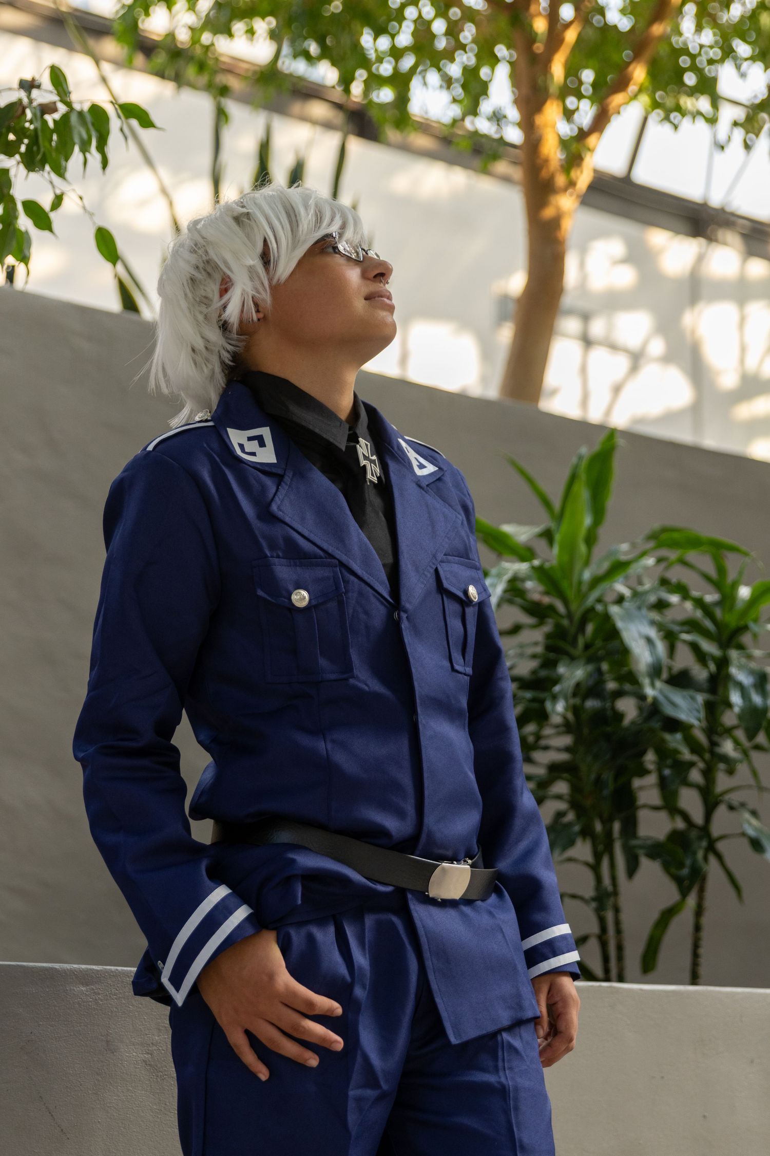 A figure wearing a navy blue uniform with striped cuffs stands against a backdrop of trees and a white wall.