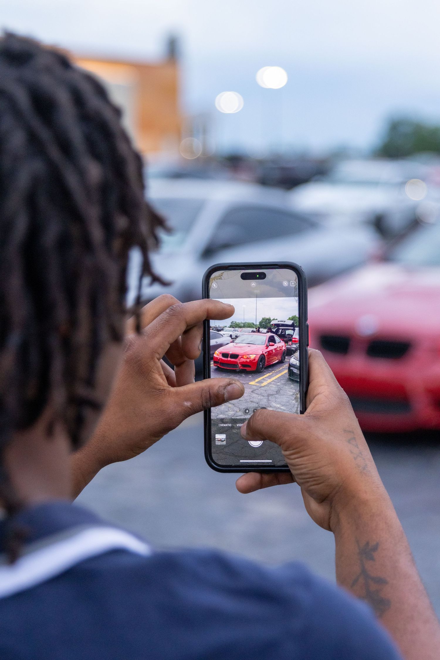 Person taking photo of red sports car with smartphone in parking lot during dusk hours.
