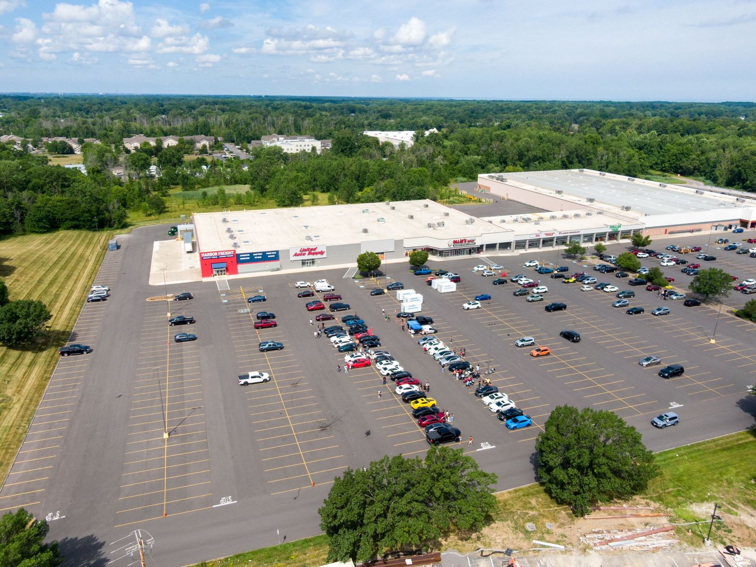 Aerial panorama of retail parking lots and storefronts surrounded by green trees with scattered cars visible.