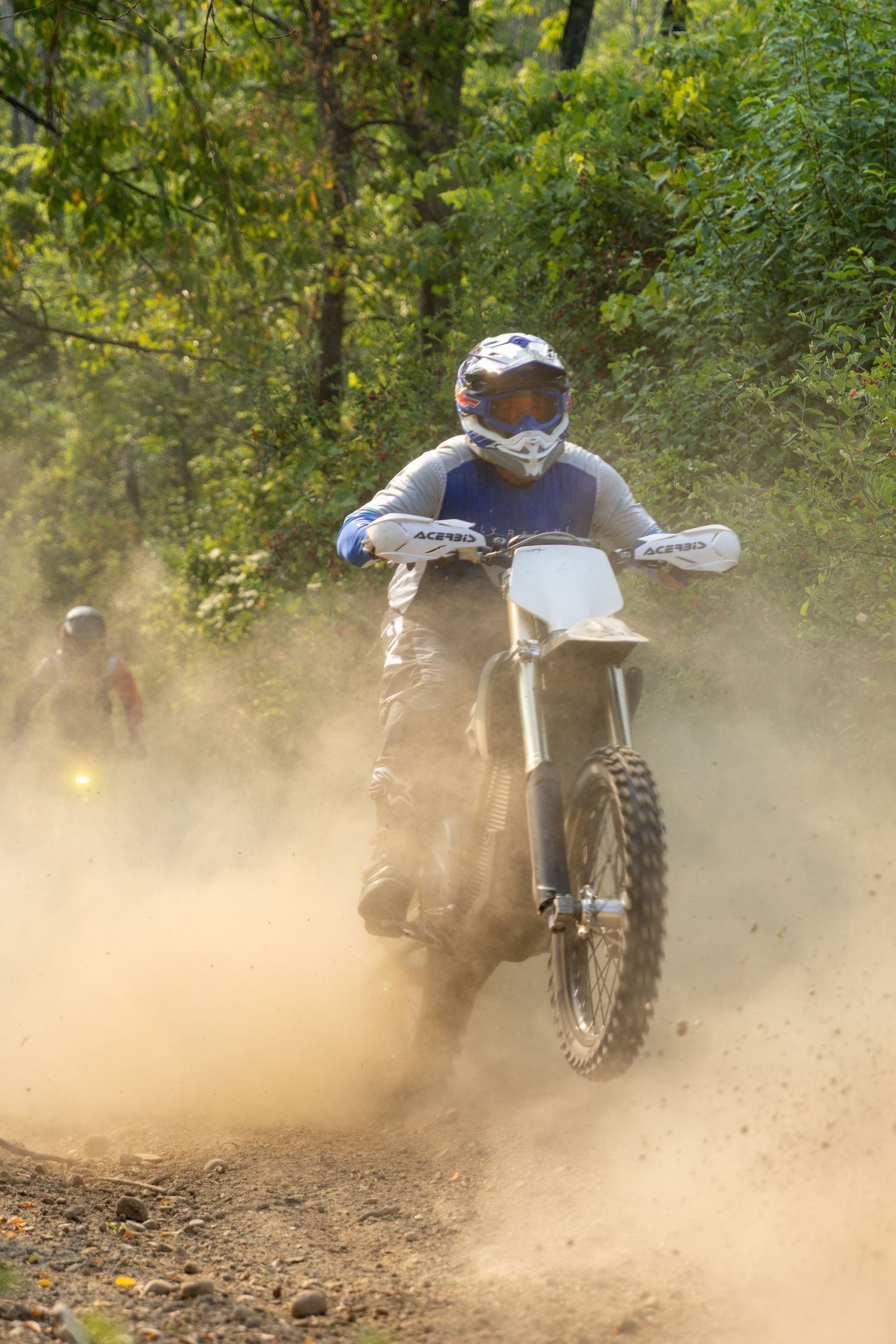 Dirt bike rider kicking up dust while racing through a wooded trail on a sunny day.
