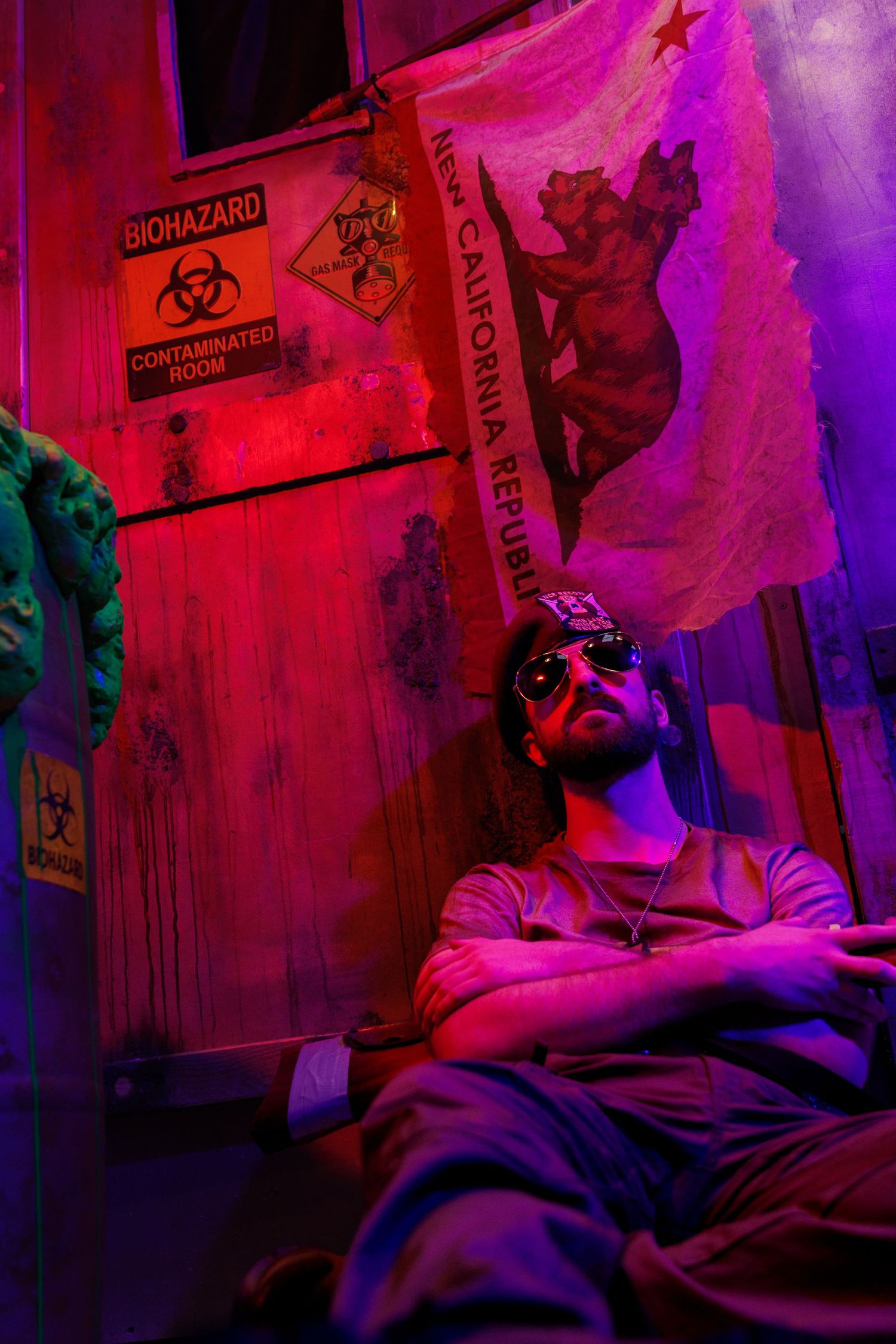 A bearded man in sunglasses sits with arms crossed under a New California Republic flag and biohazard signs in a red and purple lit room.