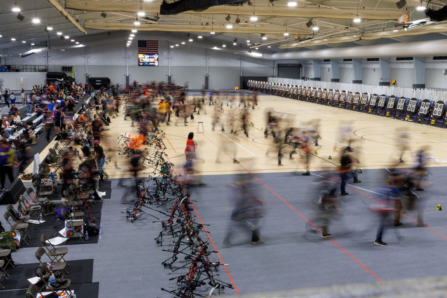 Large indoor archery competition with archers and compound bows in motion blur.