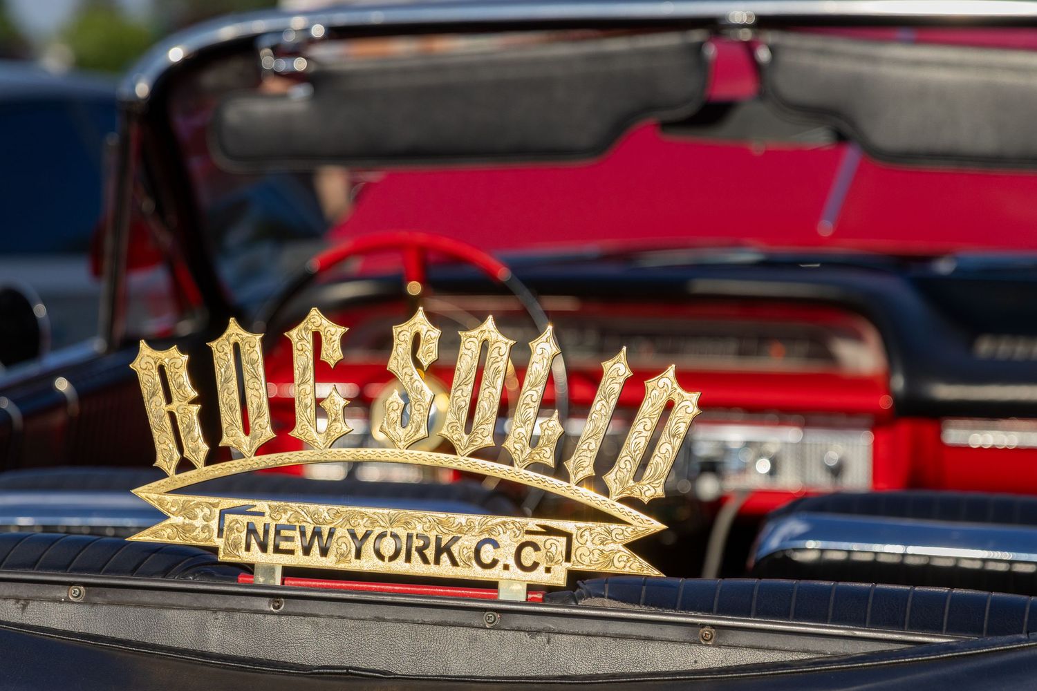 A gold 'NYC Car Club' metal badge emblem mounted on a car dashboard with a red vehicle in the background.
