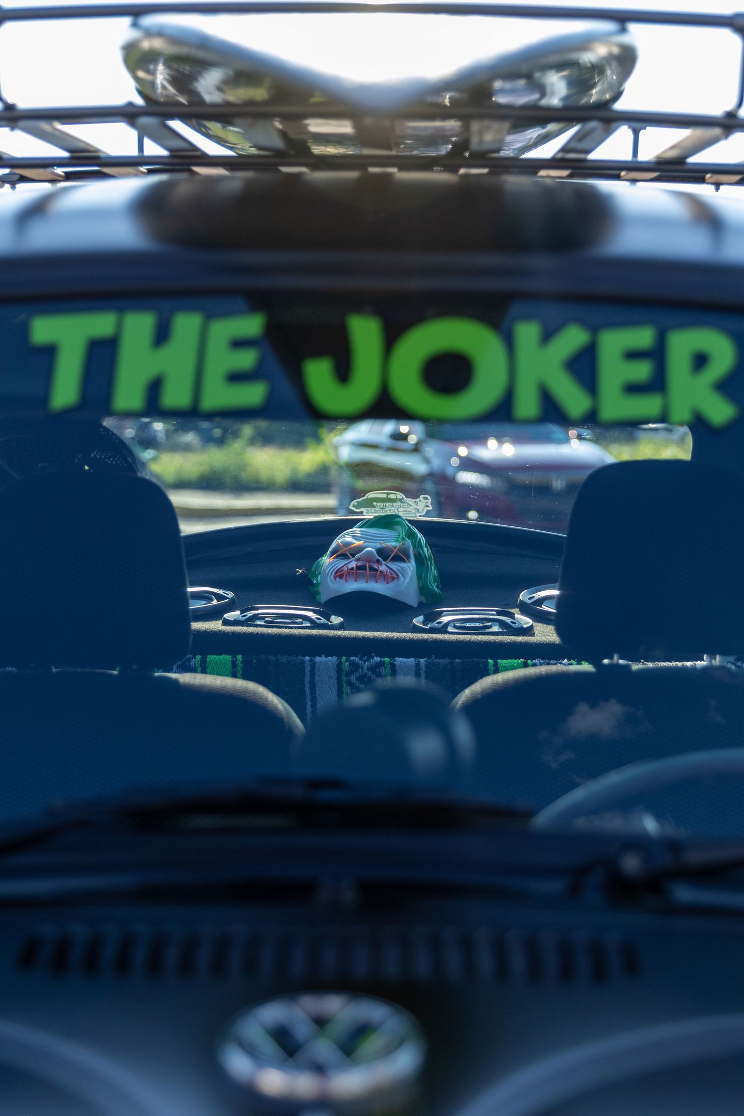 A rear window view of a parked green car with a 'The Joker' text decal visible through the glass.