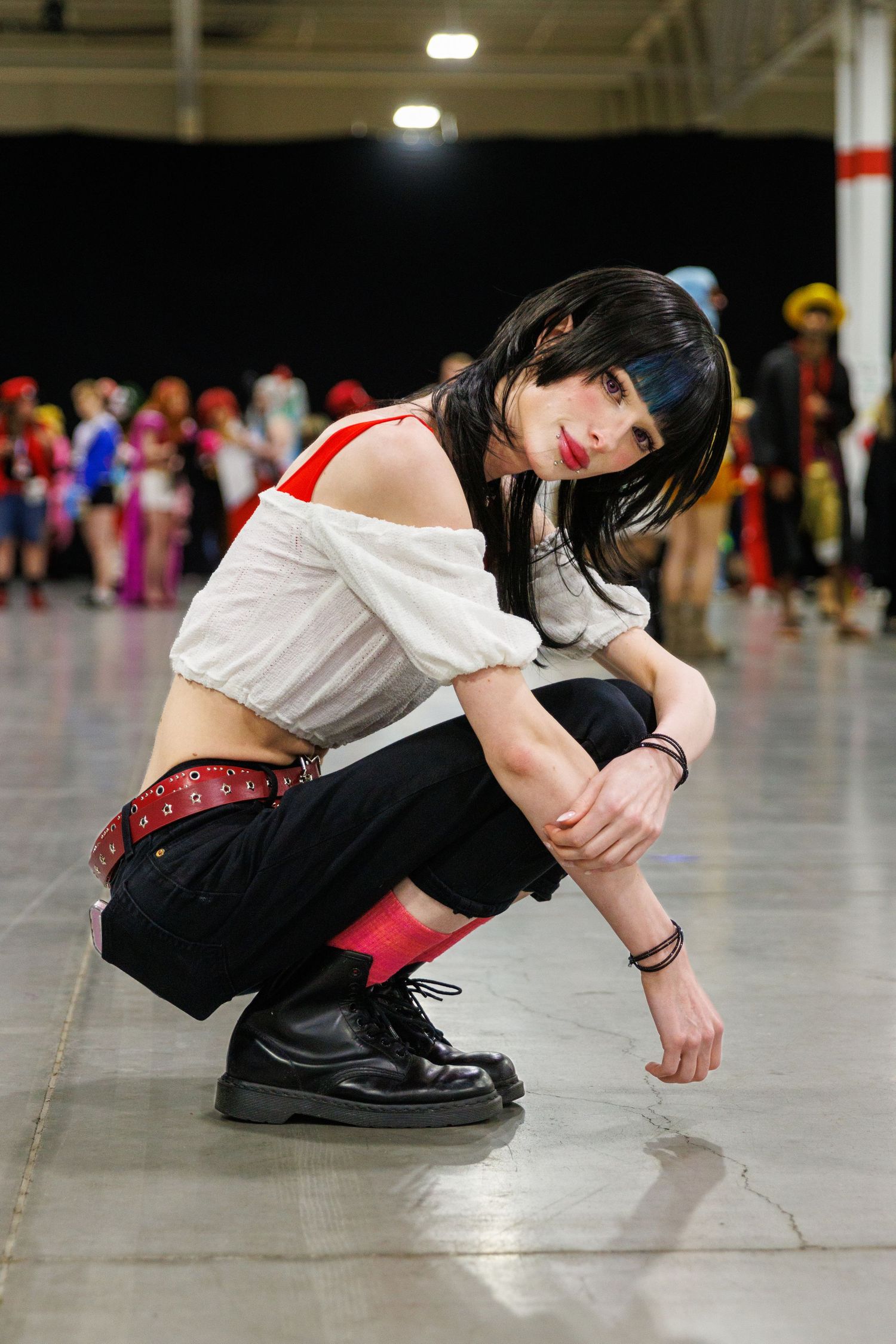 Female cosplayer in white crop top and black pants crouching on convention floor amid colorful crowd.