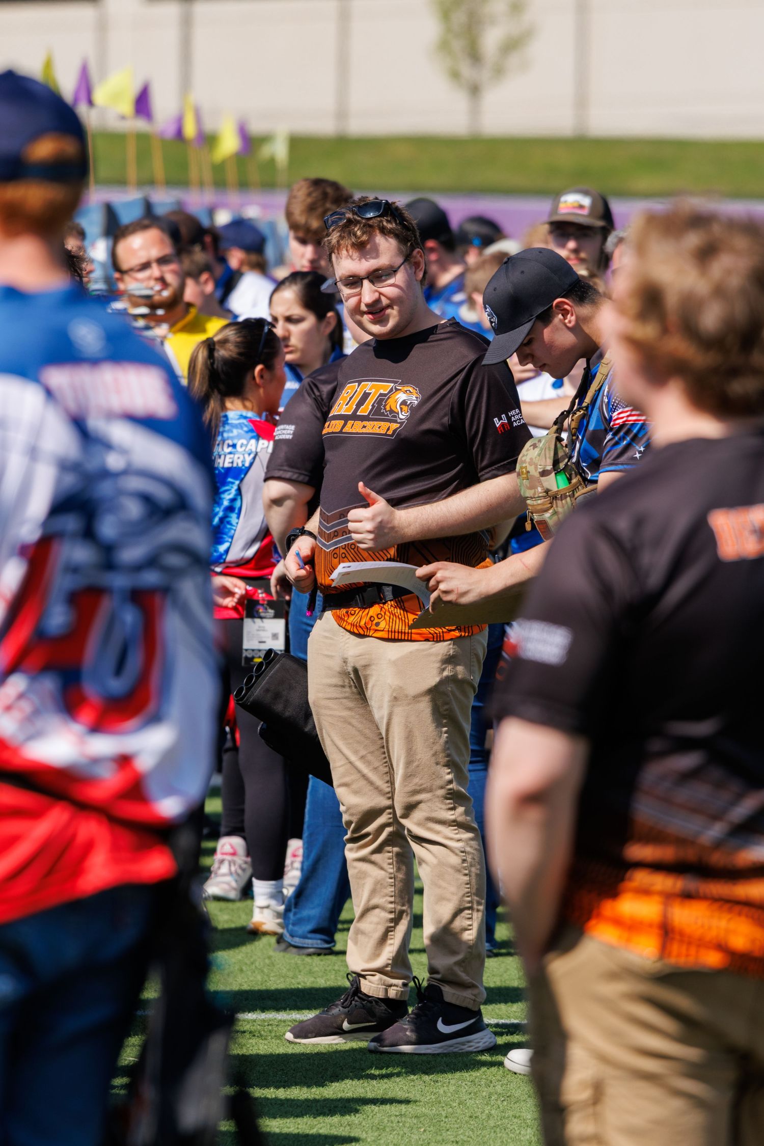 Young man in BTS shirt smiles while holding orange bow at crowded archery tournament.