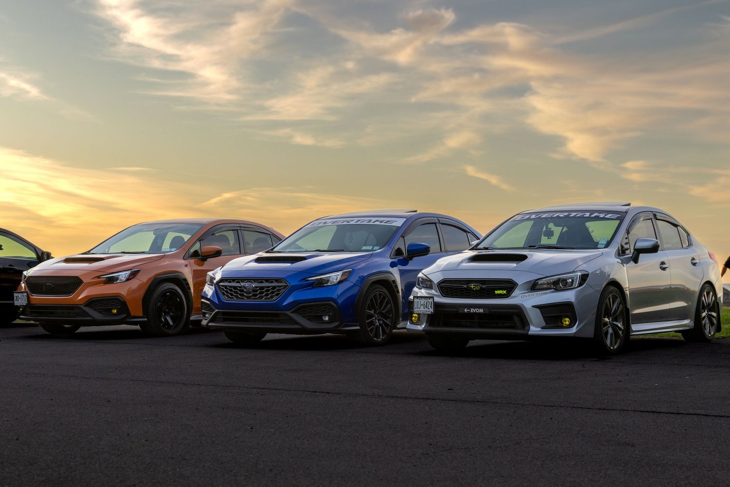 Five Subaru WRX sports cars lined up at sunset showcasing different colors and model years against a dramatic sky.