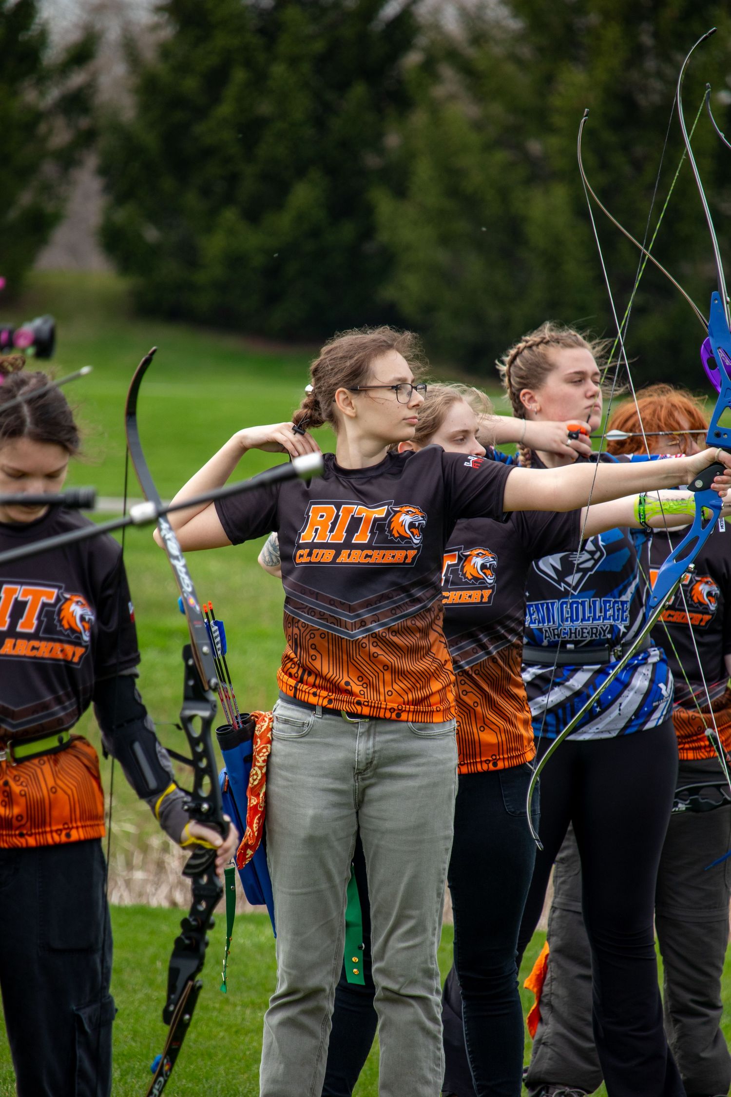 Group of archers in orange team uniforms practice with their bows on an outdoor range.