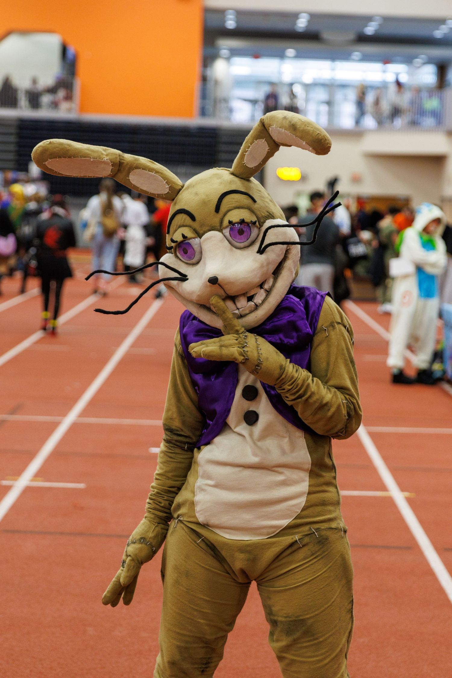 Person wearing a golden rabbit animatronic costume with purple bow tie and glasses at a cosplay convention.