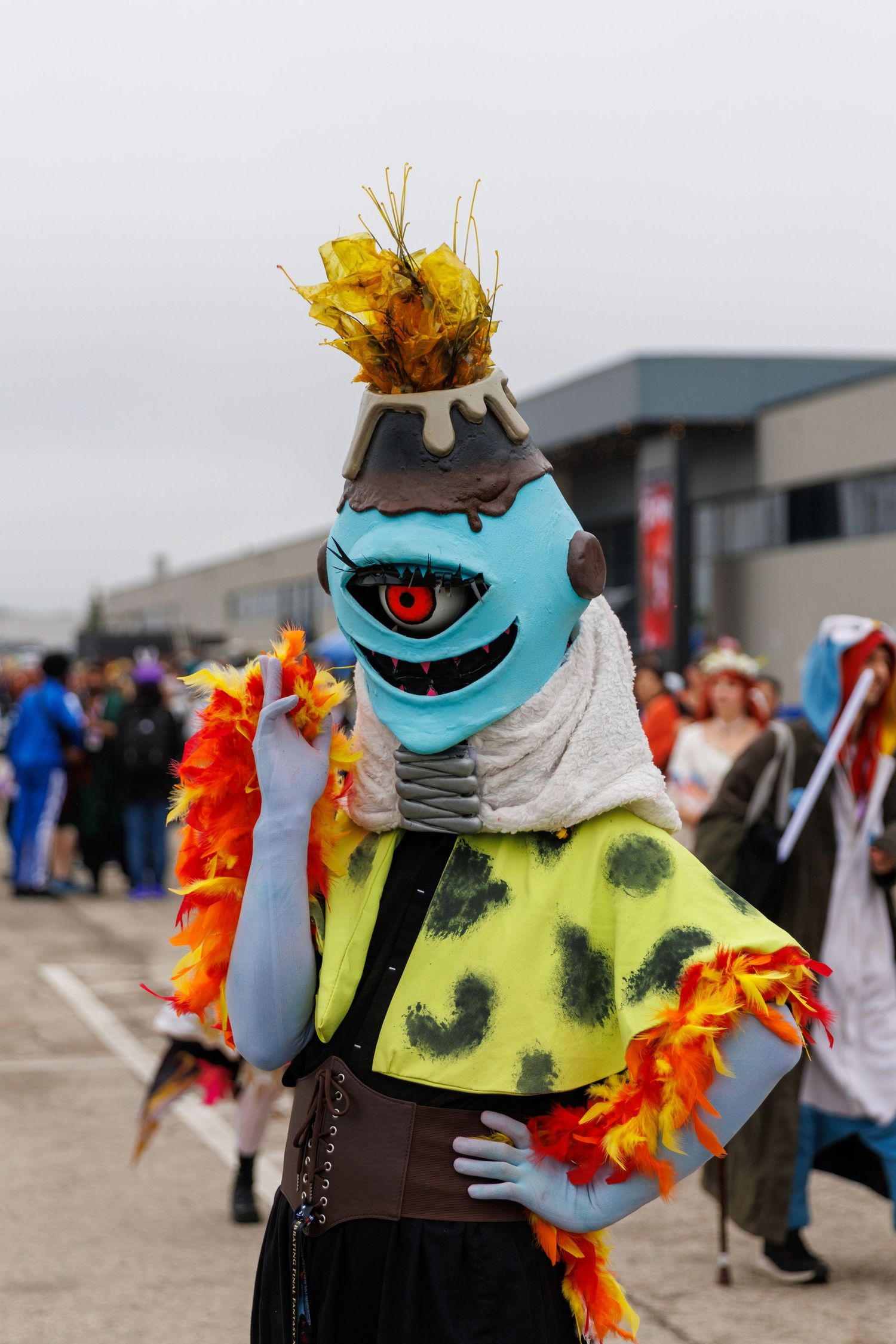 Creative cosplayer wearing a blue monster mask with one eye and colorful orange feather boa.