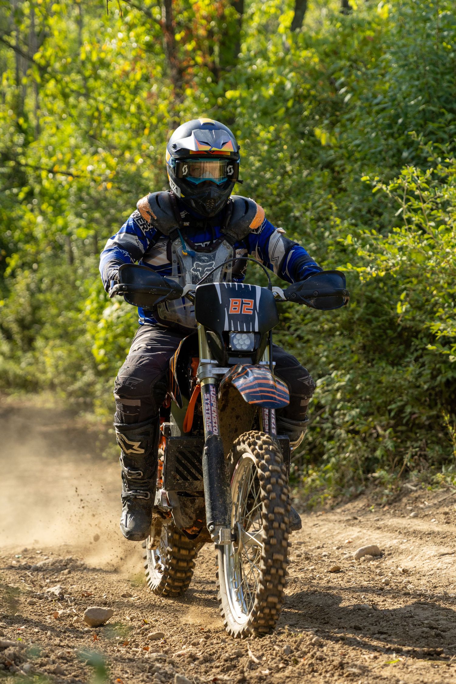 Dirt bike rider navigates a rugged trail through lush green forest terrain, kicking up dust while making a sharp turn.