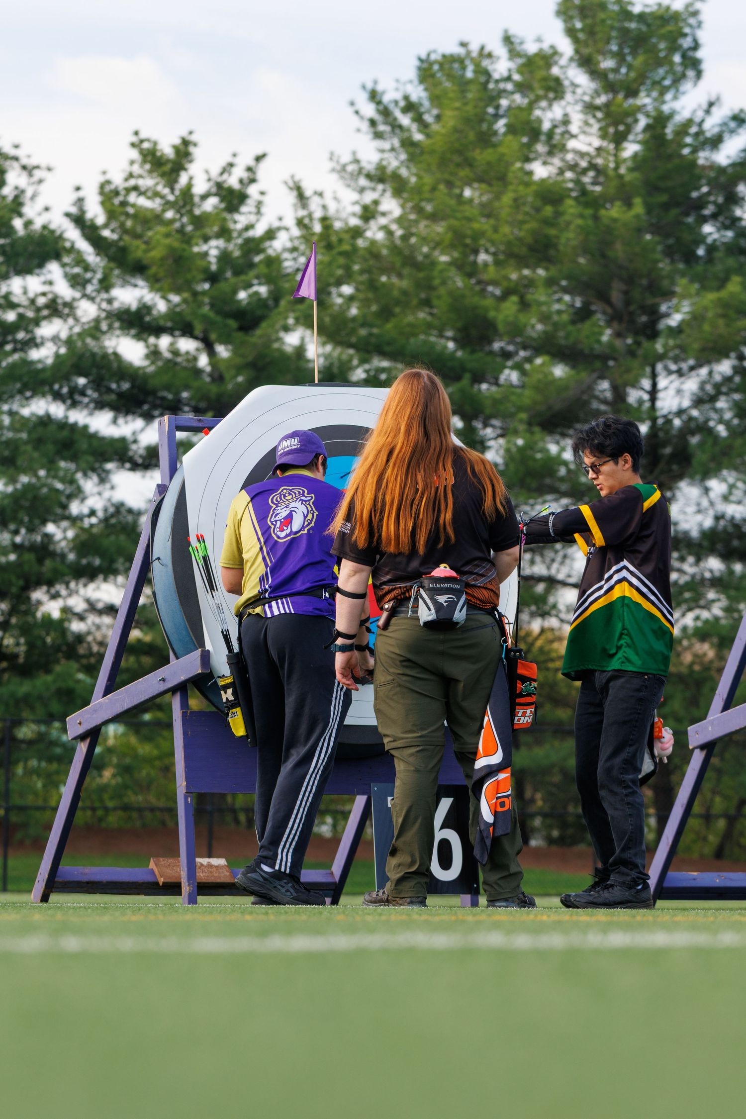 Archers gather at a target during an outdoor archery competition on a green field with trees in the background.