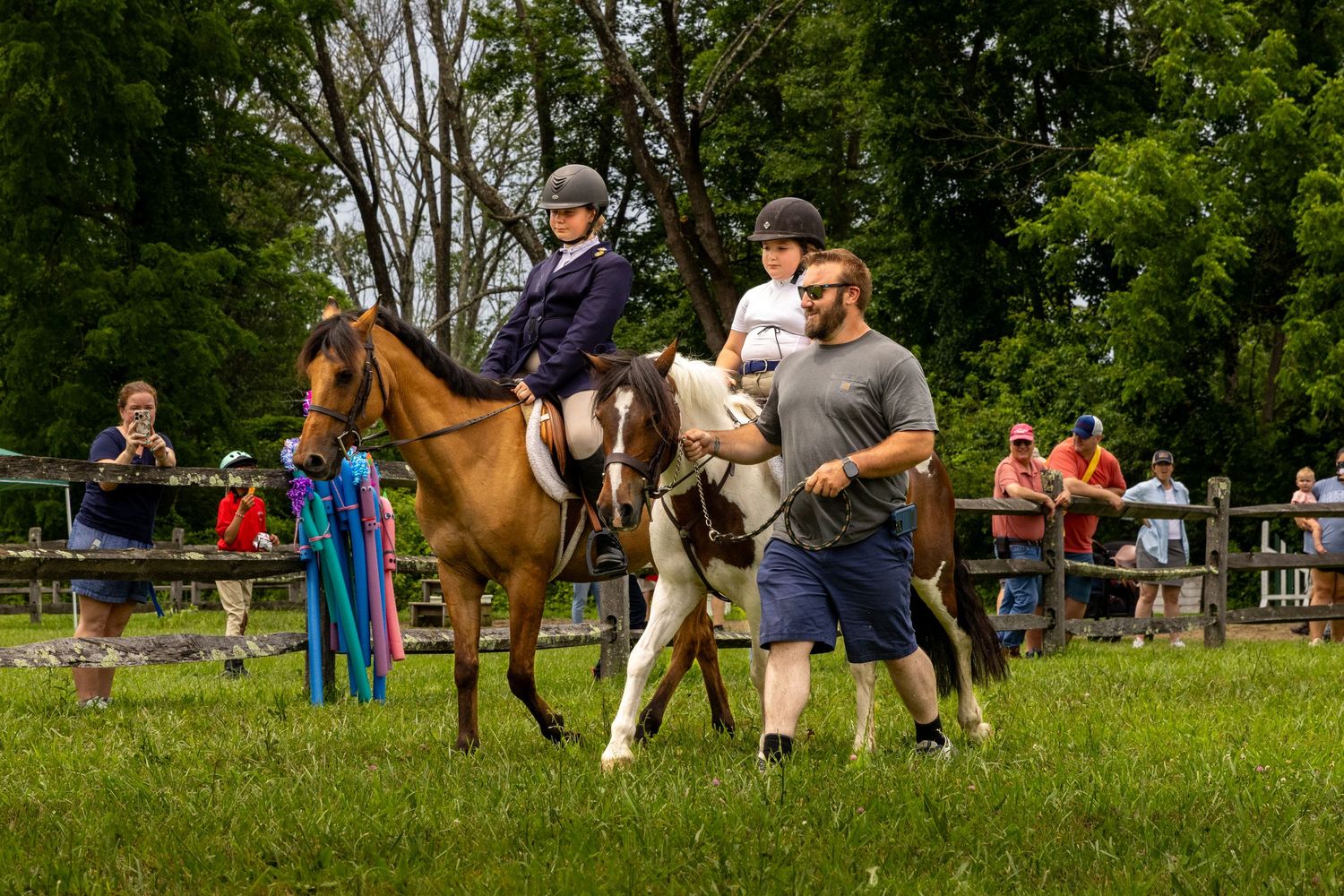 Riders and horses gather in a grassy field during an equestrian event with spectators watching.