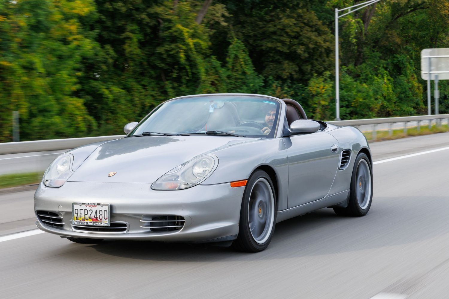 A silver Porsche 911 sports car driving on a winding mountain road through lush green forest scenery.