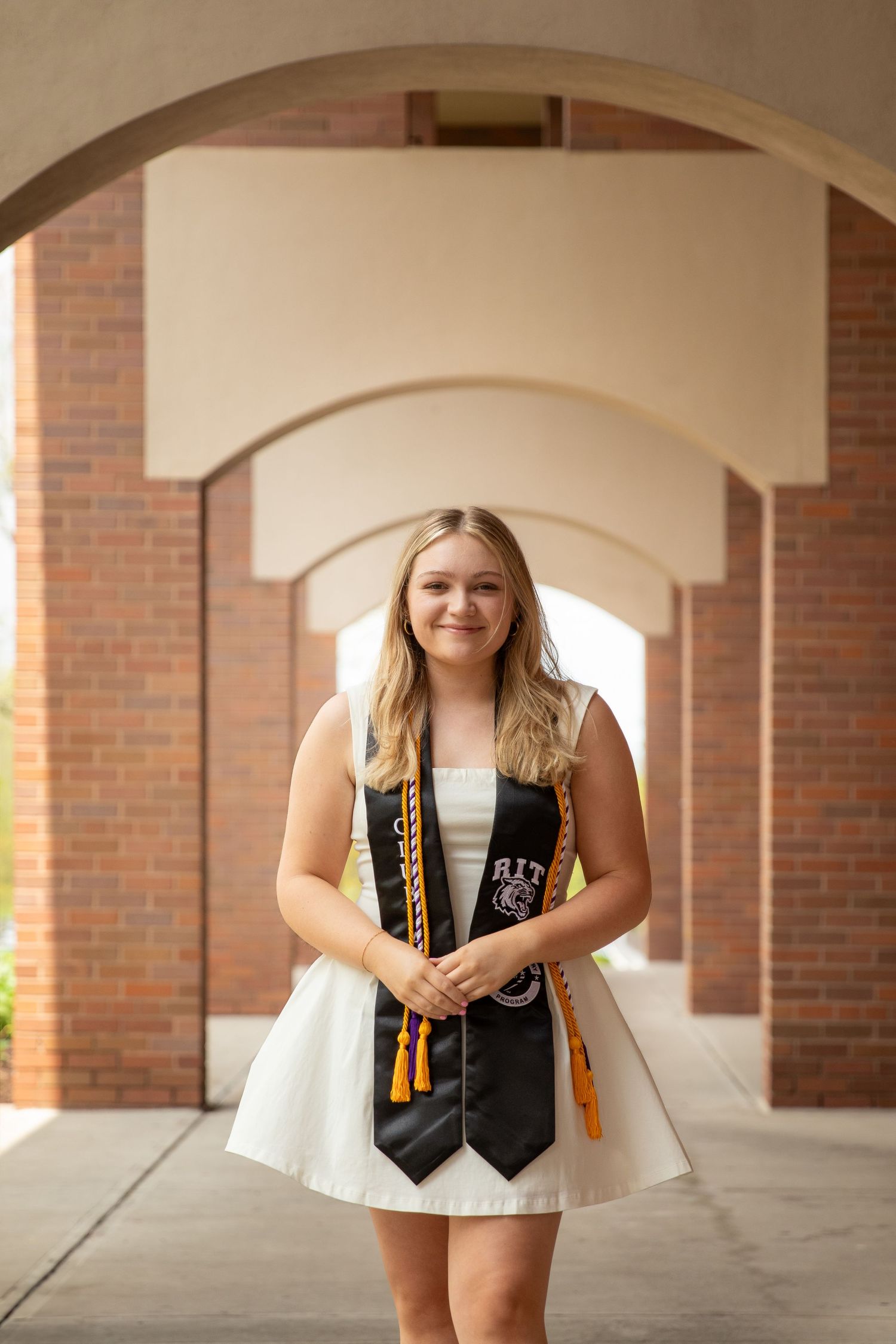 Graduate in a white dress and graduation stole stands under brick archway.