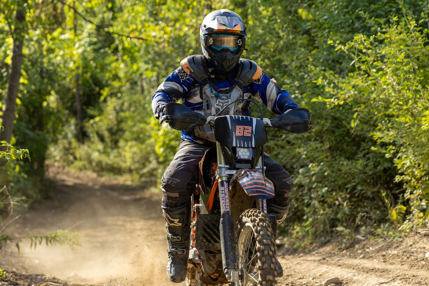 A motocross rider in blue gear and helmet races down a dusty dirt trail surrounded by lush green trees.