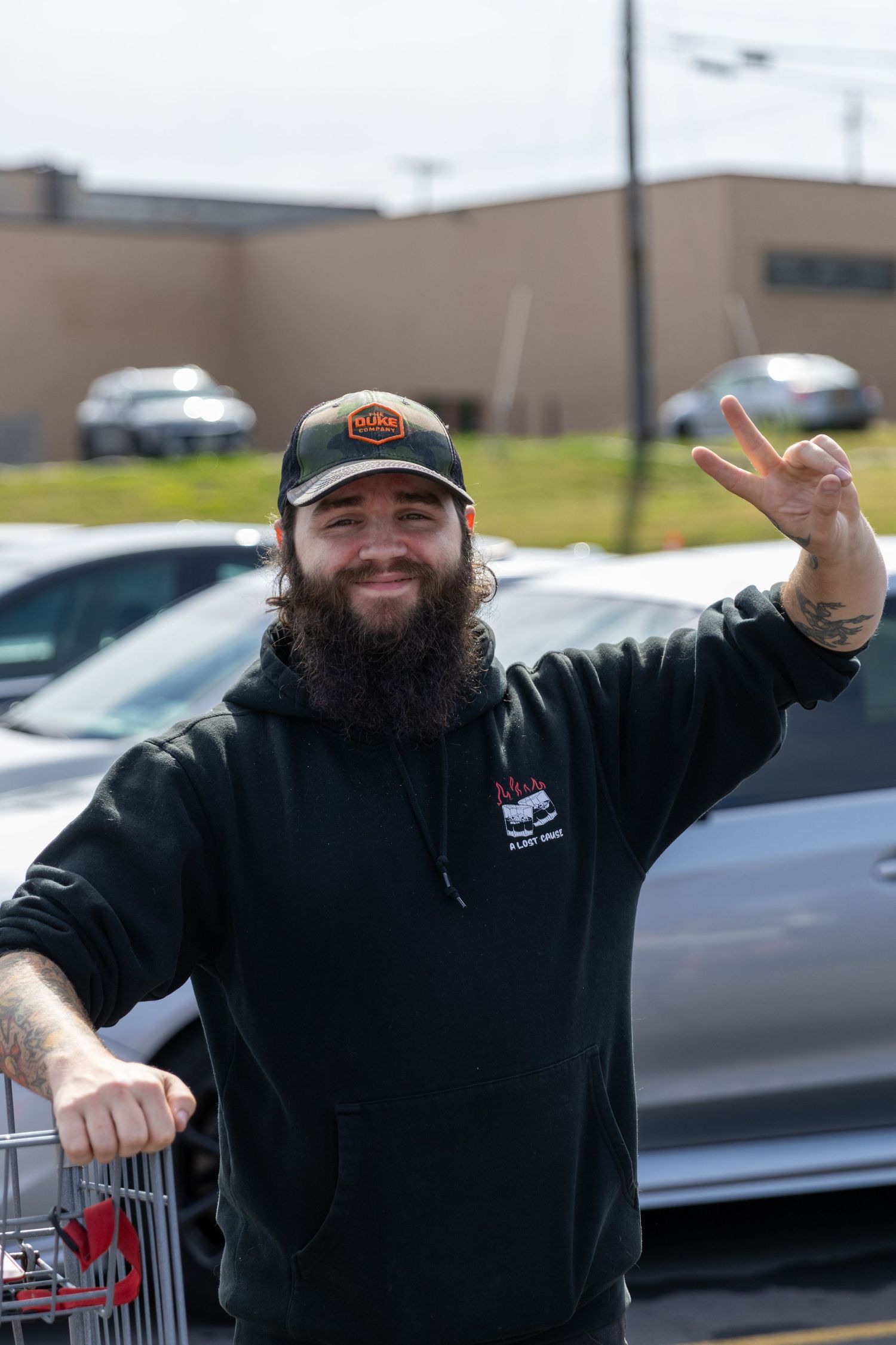 A person in a black sweatshirt making cheerful gestures outside a building with cars in the background.