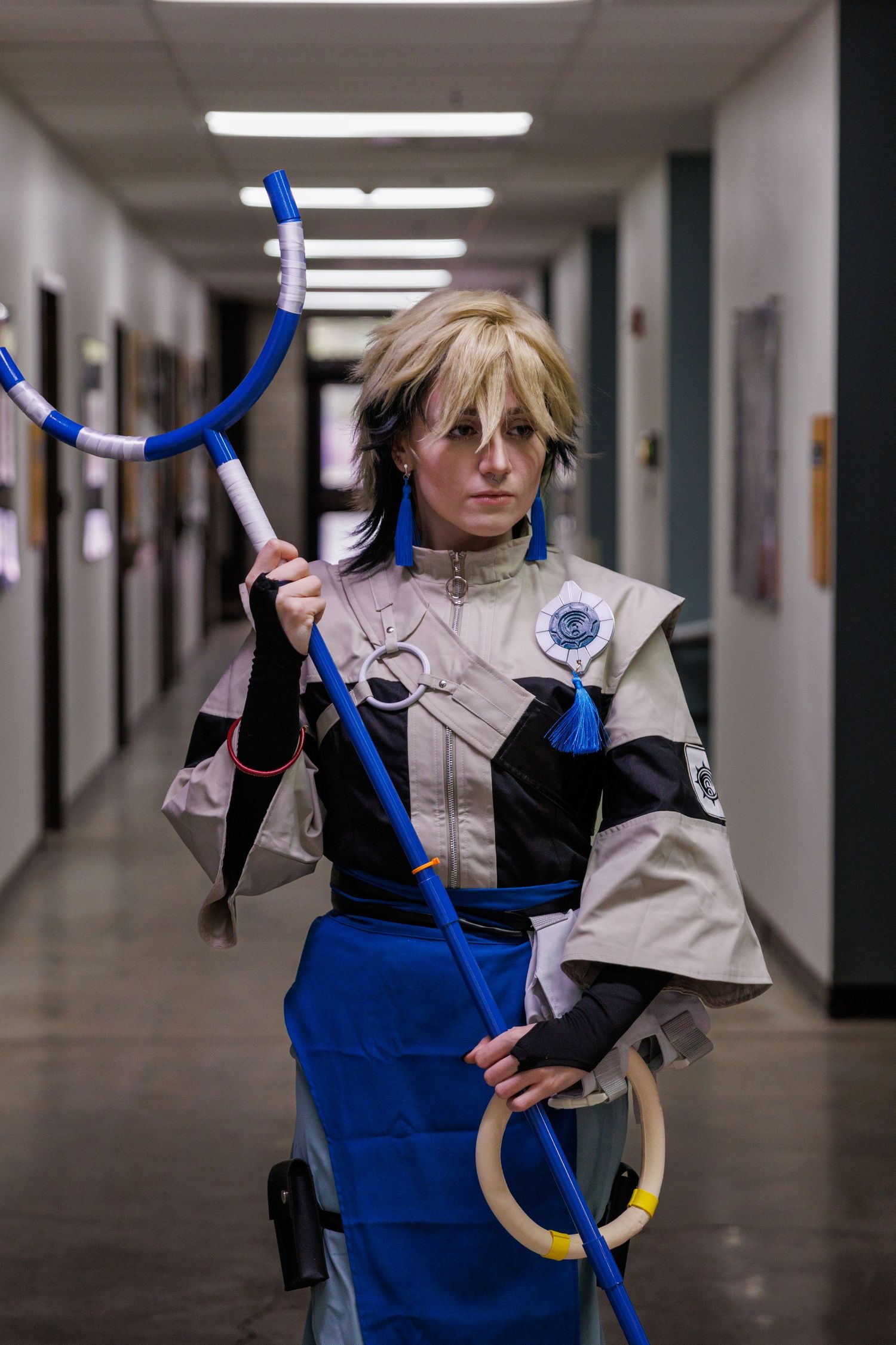 Cosplayer dressed in anime-inspired outfit holding a blue staff prop in an indoor hallway.
