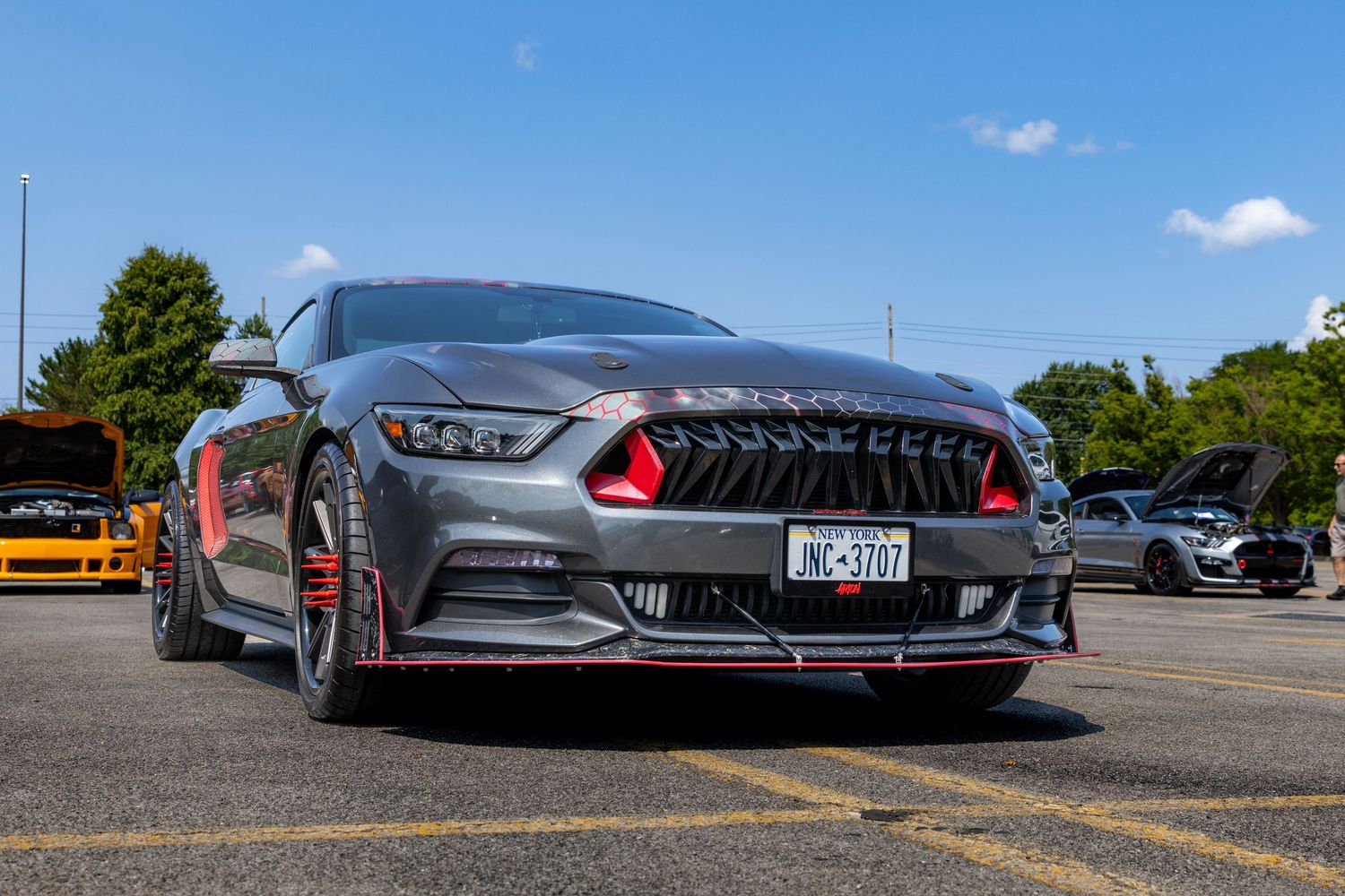 Modified Ford Mustang in metallic grey with red accents and custom front splitter parked in a sunny lot.