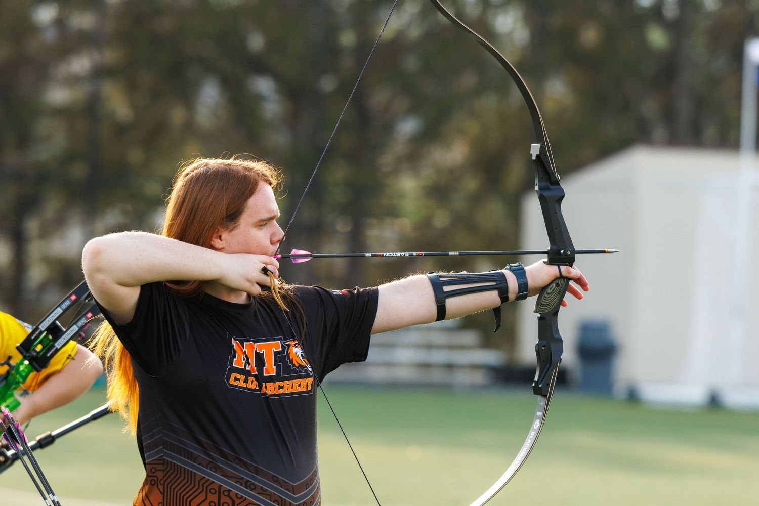 Female archer in black NT club archery shirt draws a recurve bow outdoors during a competition.