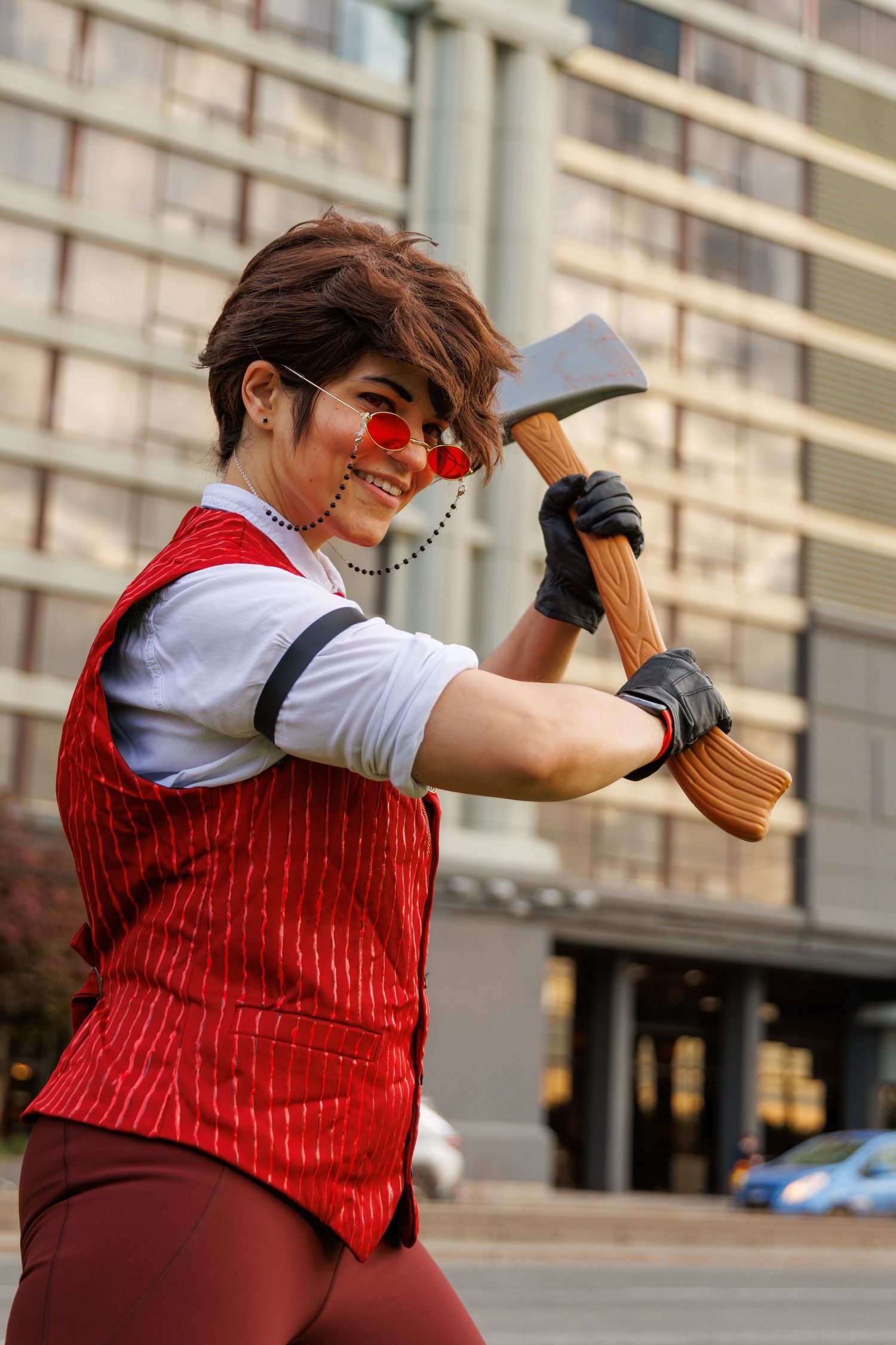Cosplayer dressed in red vest and sunglasses poses with a toy axe outdoors at a convention event.